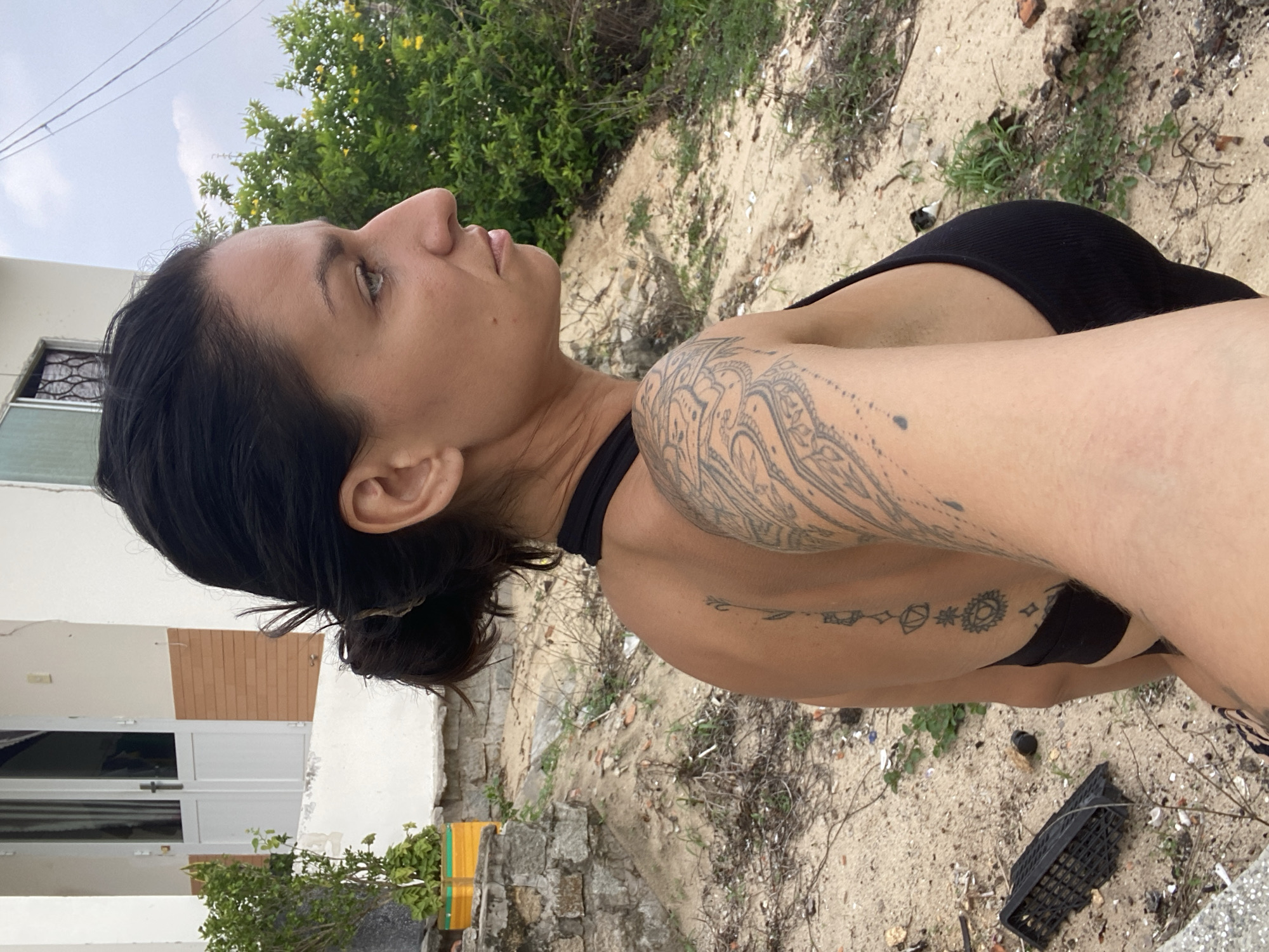 A medium shot shows a woman in profile looking to the right. She has dark hair pulled back in a bun and is wearing a black tank top. She has tattoos on her right arm and along her spine. The background is a sandy area with some greenery and a white building with orange trim. The scene appears to be outdoors, possibly in a semi-rural or coastal area. The lighting suggests it is daytime.