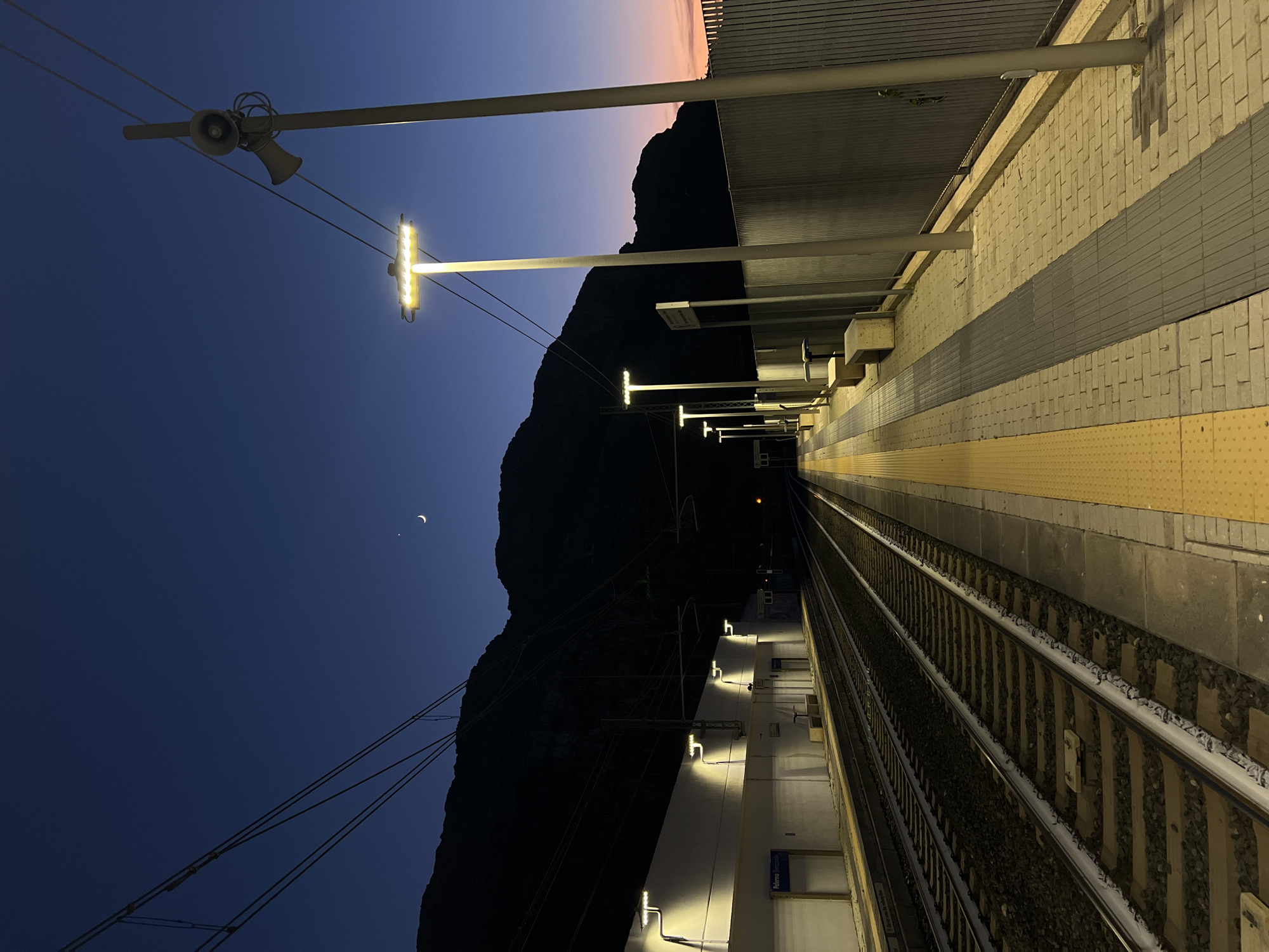 The image depicts an empty train station platform at dusk. The sky is a deep blue, transitioning to a faint orange glow on the horizon. A sliver of a crescent moon is visible in the upper portion of the sky. Mountains are silhouetted against the darkening sky. The platform is illuminated by artificial lights, casting a warm glow. Train tracks run parallel to the platform, disappearing into the distance. A fence runs along the edge of the platform, separating it from the surrounding area. There are no people visible in the image. The station appears to be located in a somewhat remote or mountainous area, given the prominence of the mountains in the background. Based on the visible signage, the station is identified as "Palermo Sferracavallo".