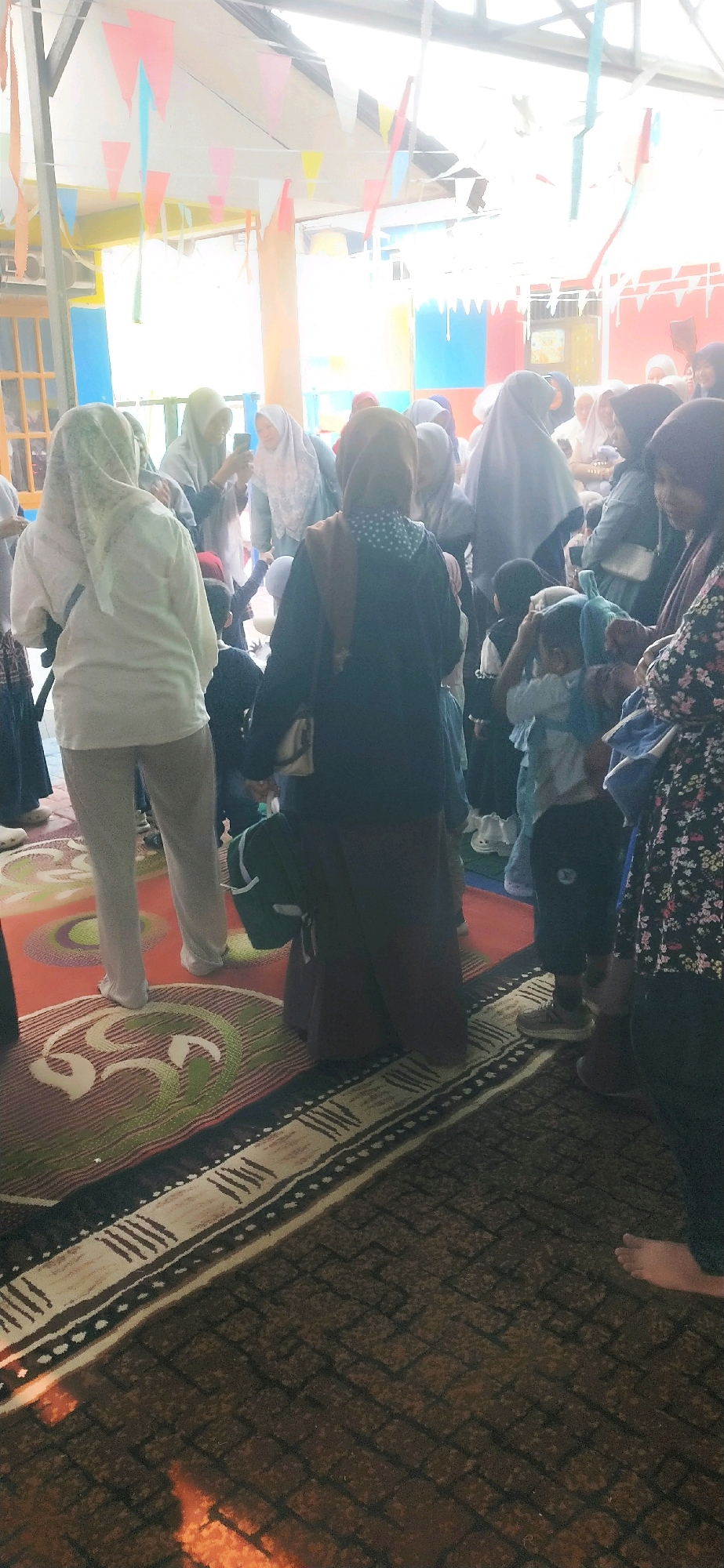 In Masukau, Indonesia, a gathering is taking place outdoors, possibly at a school or community center, under a covered area decorated with colorful triangular flags. A group of people, primarily women and children, are present. Many of the women are wearing hijabs. One woman in the foreground, wearing a white hijab and jacket, is standing with her back to the camera, looking towards a group of children. Another woman to her left is holding a smartphone, seemingly taking a picture. A child in the center is adjusting their clothing. The ground is covered with rugs, one of which has a circular pattern. The background shows colorful walls and windows, suggesting a building structure.