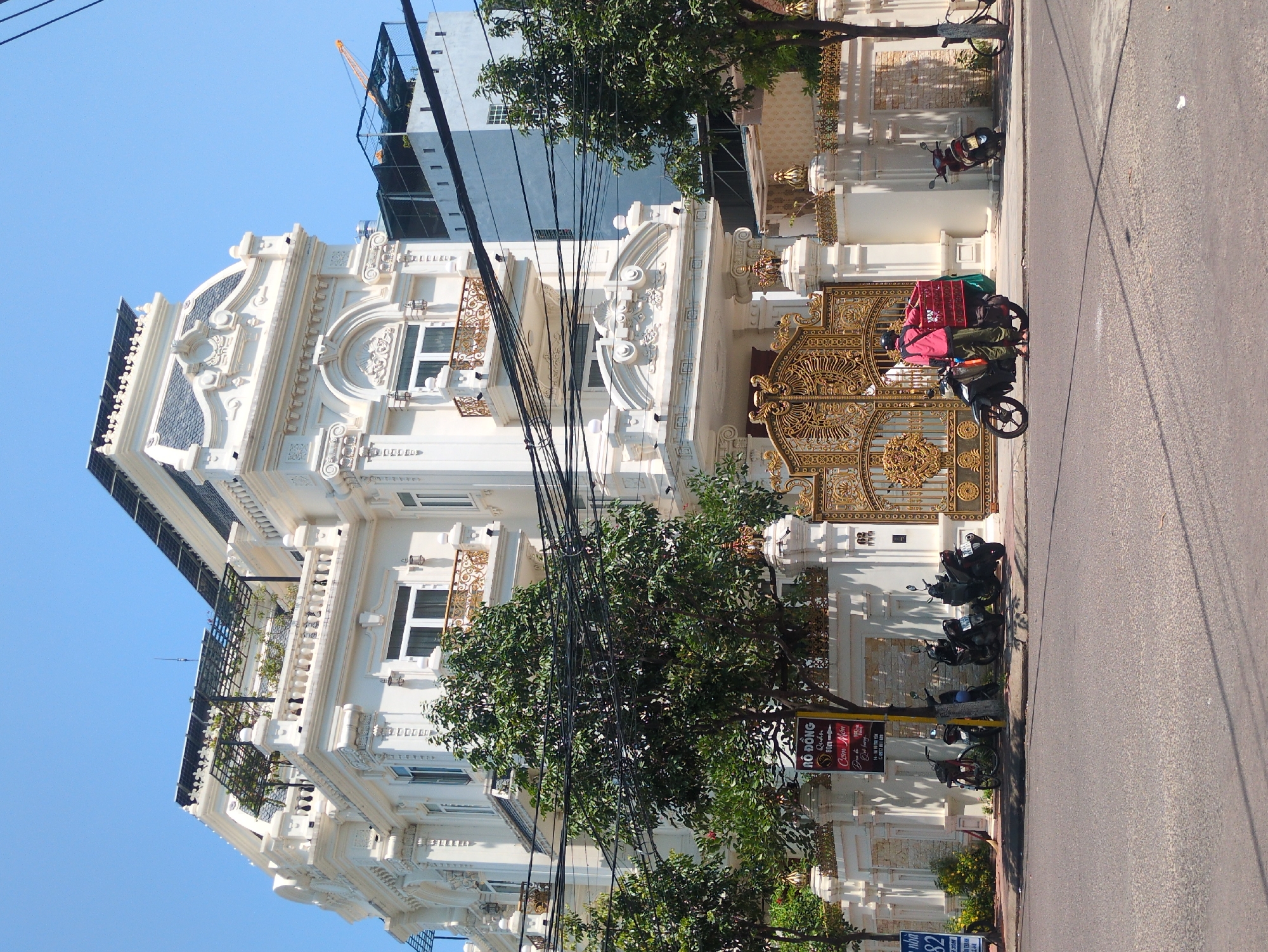 The image depicts a multi-story, ornately decorated building in Quy Nhon Ward, Vietnam. The building is primarily white with detailed architectural features. A large, ornate gold gate is prominent at the front of the property. A person on a scooter, with a red crate, is visible near the gate on the street. Several parked scooters line the curb in front of the building. A sign is near a tree on the sidewalk. Overhead power lines are visible. A modern building is behind the white building. The sky is clear and blue.
