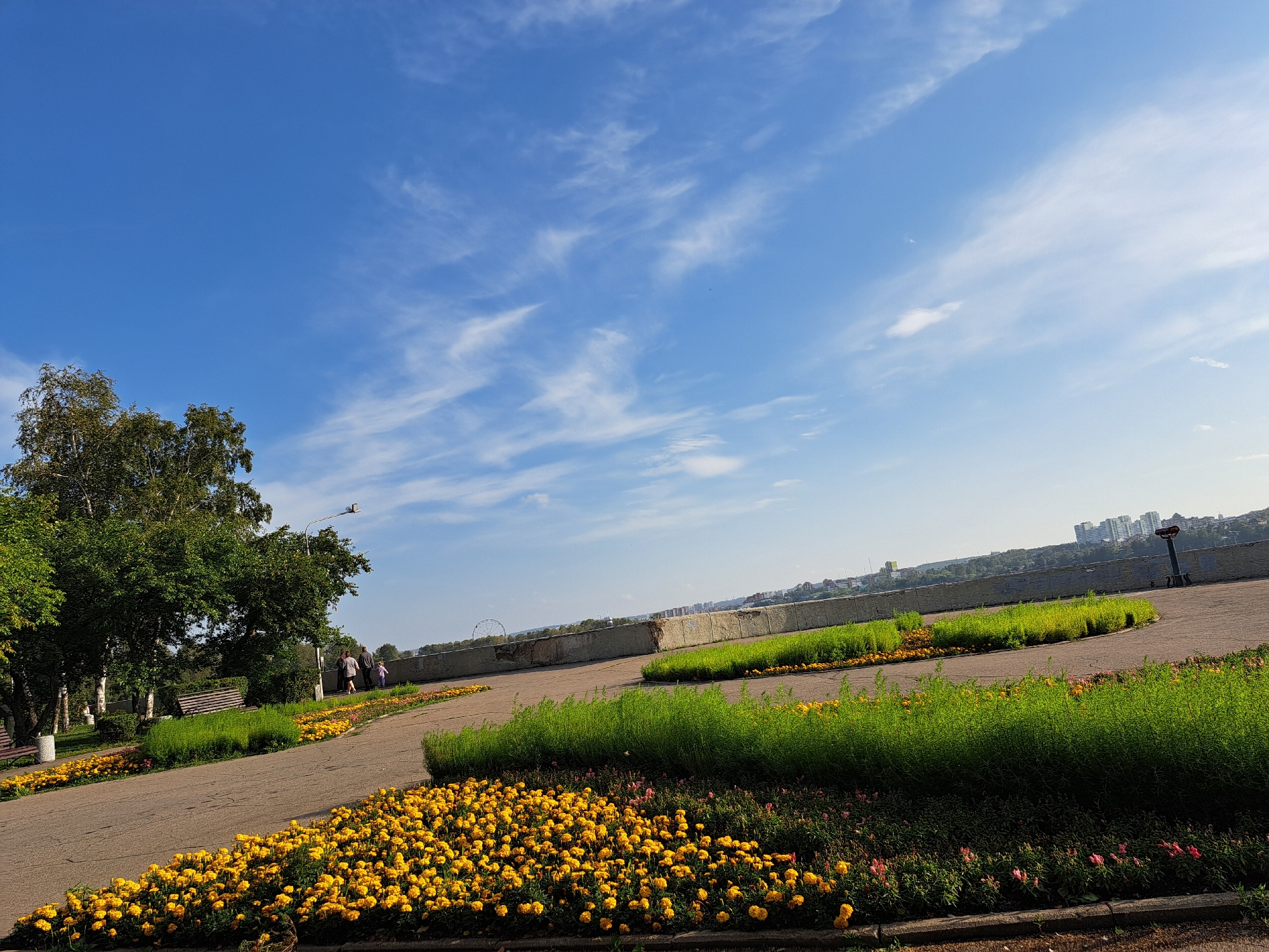 The image depicts a park scene with a paved pathway winding through flowerbeds of yellow and pink blooms. Tall green bushes border the pathway, which leads towards a city skyline in the distance. A concrete wall runs along the edge of the park, offering a view of the urban landscape. Trees with green foliage are visible on the left side of the image, and a park bench is nestled amongst them. A few people are strolling along the pathway. The sky is a clear blue with scattered white clouds. The scene is consistent with the Upper Embankment of the Angara River in Irkutsk, Russia, a popular recreational area offering views of the city and the river.