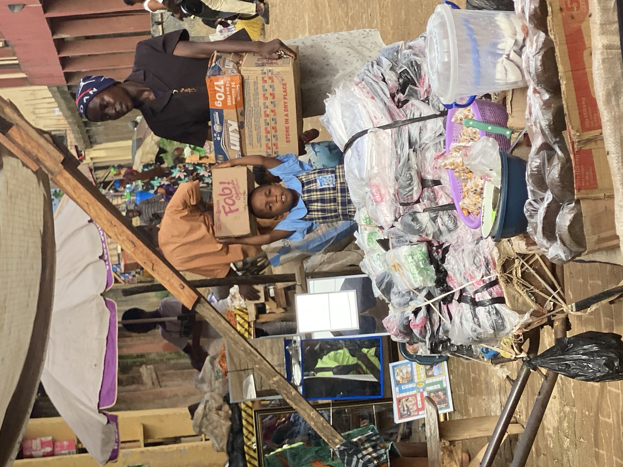 The scene is a market in Kafanchan B, Nigeria. An adult and a child are carrying cardboard boxes on their heads. The child wears a blue and white uniform. The adult wears a dark shirt and a blue hat. Stacks of goods wrapped in plastic bags surround them. The market stall includes items such as a white bucket, a blue bowl, and a purple basket, along with framed pictures and mirrors. The background shows a crowded marketplace with various structures.