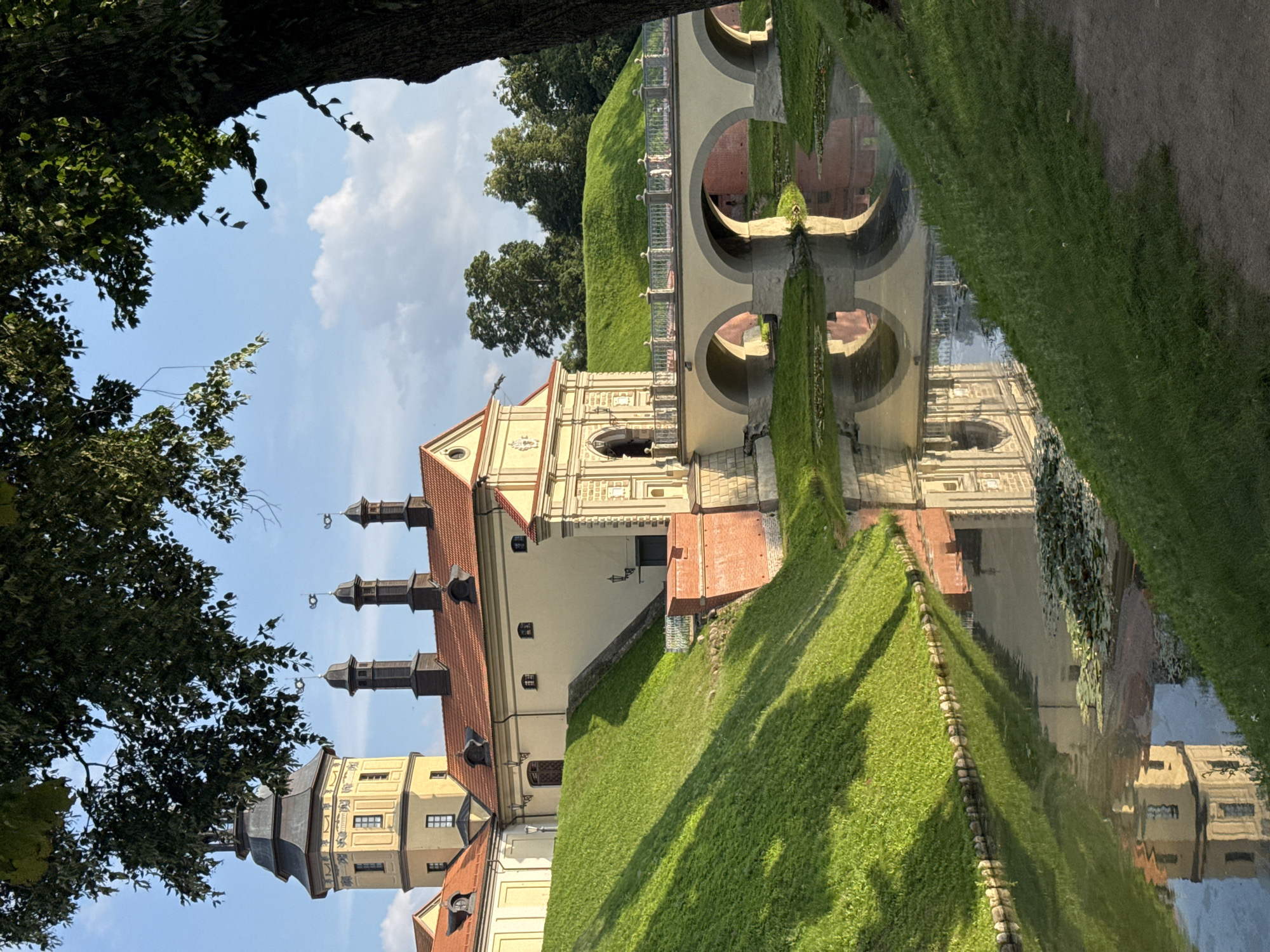 The image depicts a view of Nesvizh Castle in Nesvizh, Belarus. The castle is a large, multi-towered structure with a yellow facade and red tiled roofs. A prominent tower with a black spire rises on the left side of the image. In front of the castle, a wide moat is visible, reflecting the castle's architecture and the surrounding green landscape. A stone bridge with arched openings spans the moat, connecting the castle grounds to the foreground. The banks of the moat are covered in lush green grass, with shadows cast by trees. The sky is blue with scattered white clouds, and a large tree with dark bark and green leaves occupies the right side of the frame, partially obscuring the sky and the castle. The overall scene suggests a sunny day at a historic landmark.