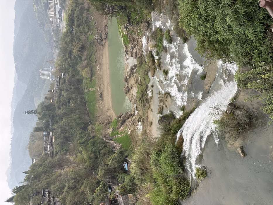 The image captures an elevated, wide view of Furong, China, under an overcast sky. In the immediate foreground, a broad waterfall cascades white, turbulent water over dark rock formations from left to right, emptying into a series of smaller rapids and a calmer, darker stream. Dense green vegetation, including tall grasses and bushes, borders the water.
The midground features the river widening into a greenish-blue, relatively still pool. The banks of this pool are exposed, revealing light brown, muddy-rocky terrain interspersed with patches of green grass, indicative of low water levels. The steep hillsides on both sides of the river are densely covered with varying shades of green foliage and trees. On the left hillside, multiple traditional-style buildings with dark roofs are constructed into the terrain, connected by visible pathways and stairs. A distinctive small, blue-roofed structure is visible on the left bank near the waterfall's edge.
The background shows rolling, forested mountains stretching across the horizon. A modern, multi-story white high-rise building stands prominently in the valley, accompanied by several other distant structures and a faint bridge, signifying human development amidst the natural mountainous environment. No individuals are visible.