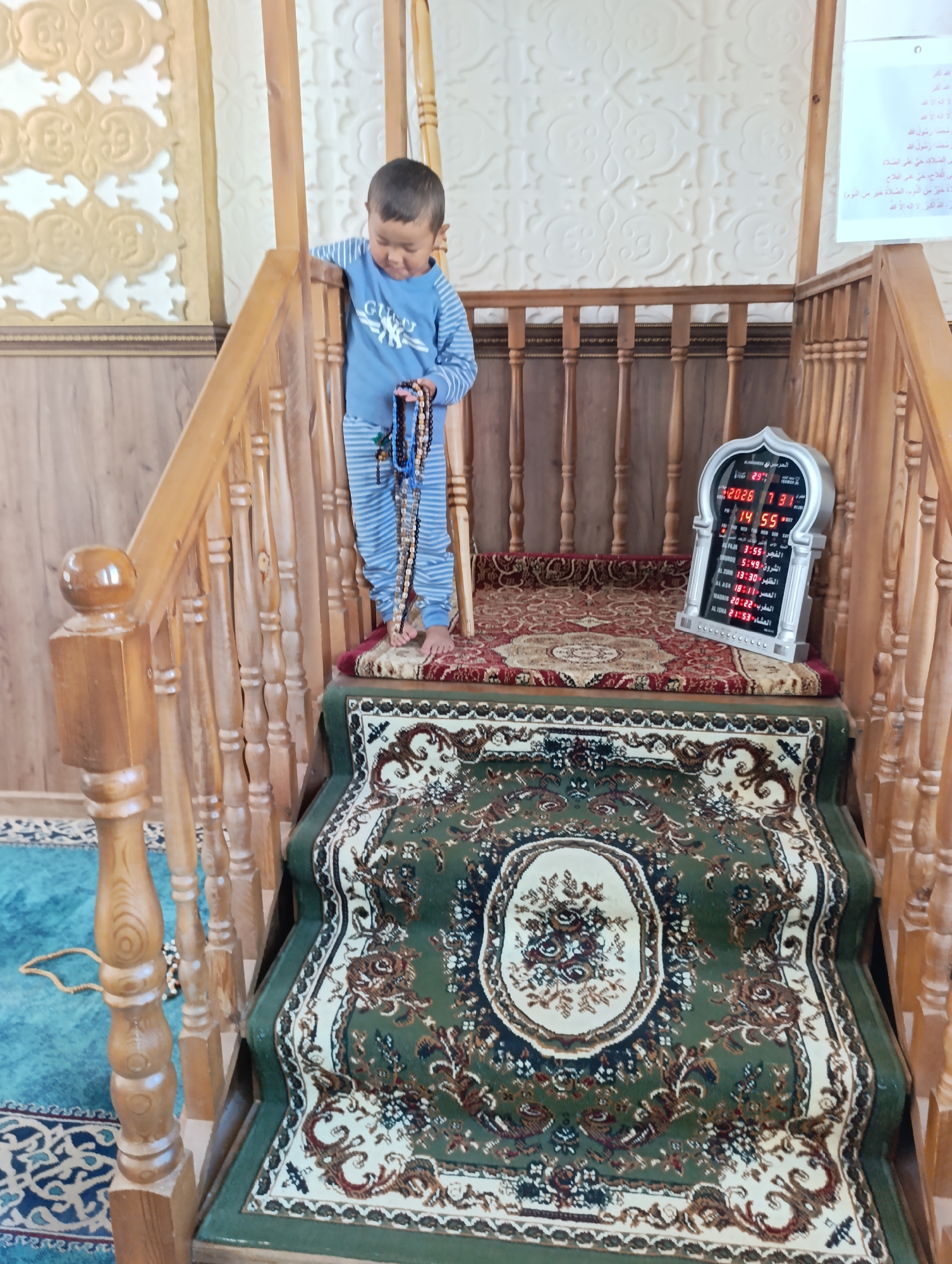 A young boy, wearing a blue and white striped long-sleeved shirt and matching pants, stands on a wooden staircase inside a mosque in Naryn City, Kyrgyzstan. He is holding prayer beads in his hands and looking down at them. The staircase is adorned with a red and gold patterned rug. To the right of the boy, on a lower step, is a digital clock displaying prayer times. The mosque has wooden railings and walls, and a large, ornate carpet with green, white, and dark red patterns covers the steps. The background wall has a decorative embossed pattern. A blue carpet with a repeating geometric pattern is visible on the floor to the left of the staircase.