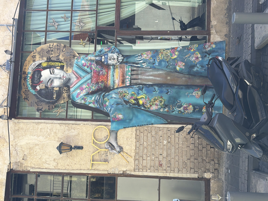 The scene depicts an urban streetscape in Tel-Aviv, Israel, featuring the facade of a building. Dominating the right side of the image is a large, multi-panel cutout artwork of a Japanese woman, styled as a geisha, affixed to the building. She has pale skin, prominent eyes, red lips, and an elaborate dark updo hairstyle adorned with yellow and white elements. Her kimono is predominantly teal and light blue, intricately decorated with multi-colored floral patterns, butterflies, and a visible dragon motif on her left sleeve (viewer's left). Her left hand holds a pair of yellow chopsticks, with the 'O' of the adjacent "TYO" sign integrated into her grip. Her right hand holds a white bottle displaying blue Japanese characters. Behind her head, visible through a multi-paned window, is a circular golden-brown panel with black Japanese text. Several golden butterfly cutouts are adhered to the window pane.
To the left of the artwork, the building facade consists of exposed brickwork below and rough, light-colored plaster above. An antique-style black lantern is mounted on the plaster. Above the lantern and to the left of the artwork, the yellow uppercase letters "TYO" are affixed horizontally to the plaster. A dark horizontal structure, possibly an awning or beam, runs above the windows, with two pigeons perched on it.
At street level, in the lower center, a dark grey or black Yamaha scooter is parked on the paved sidewalk, partially obscuring the lower section of the artwork. To the far left, a clear stemmed wine glass containing a pale yellow liquid rests atop a grey cylindrical bollard. Another grey bollard is visible to the right of the scooter. The ground is paved with light grey rectangular stones. A faint reflection of the scooter is visible in the lower right window pane.
