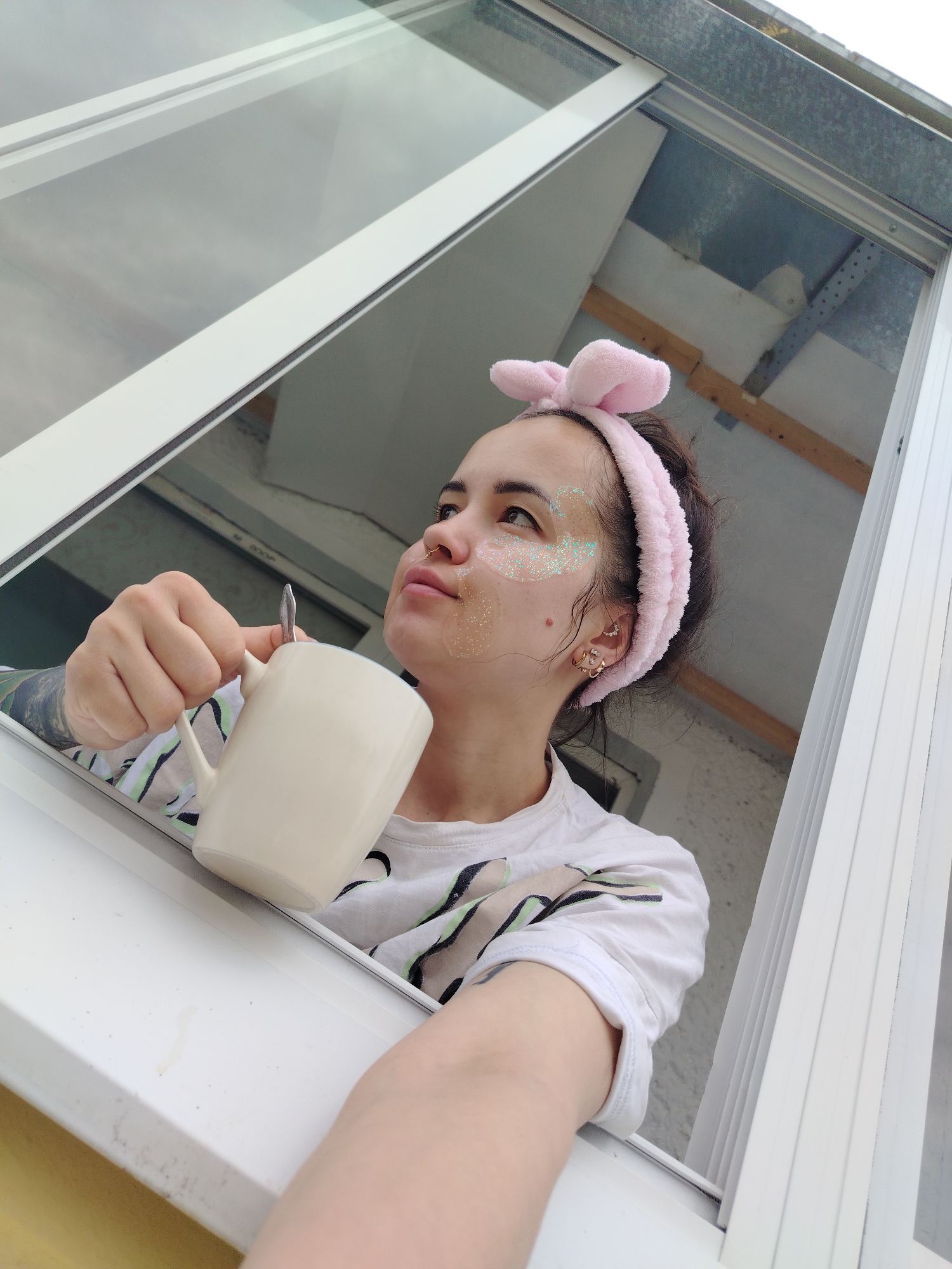 A young woman is captured in a close-up, angled shot, looking out of a white-framed window. She holds a cream-colored mug with a spoon in it, resting her arm on the window sill. Her gaze is directed upwards and outwards, suggesting a contemplative or observant mood. She wears a soft pink, plush headband with bunny ears, pushing her dark hair back from her face. Glitter is applied around her eyes and on her cheekbones, indicating she might be wearing a face mask or glitter makeup. Her attire consists of a white t-shirt with a subtle abstract pattern in light green and black. Visible accessories include gold hoop earrings and a nose piercing. A tattoo is partially visible on her left arm.
The setting appears to be an interior looking out, possibly from an upper floor or attic window, common in residential buildings in places like Noviy, Russia. The exterior view through the window shows a corrugated metal roof section, some wooden beams, and a plain white wall, suggesting a utilitarian or unfinished architectural style outside. The lighting is soft and diffused, indicating an overcast day or indirect light. The window frame itself is clean and modern, likely made of PVC. No discernible text is visible on any objects or surfaces. The overall impression is one of a quiet moment of self-care or reflection.