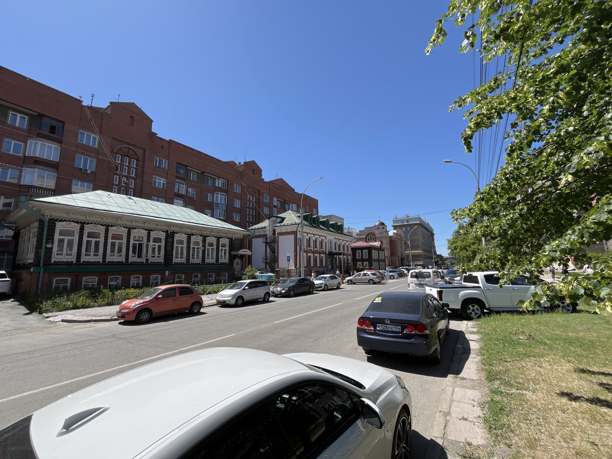 The image shows a street scene with cars parked and driving on a sunny day. A white car is in the foreground, partially obscuring a street and sidewalk. Further down the street, several other cars are visible, including a red hatchback, a dark sedan, a white pickup truck, and a white van. Buildings line the street, with a mix of brick and wooden structures. Trees with green foliage are present, with one tree's branches extending into the frame on the right. The sky is clear and blue.