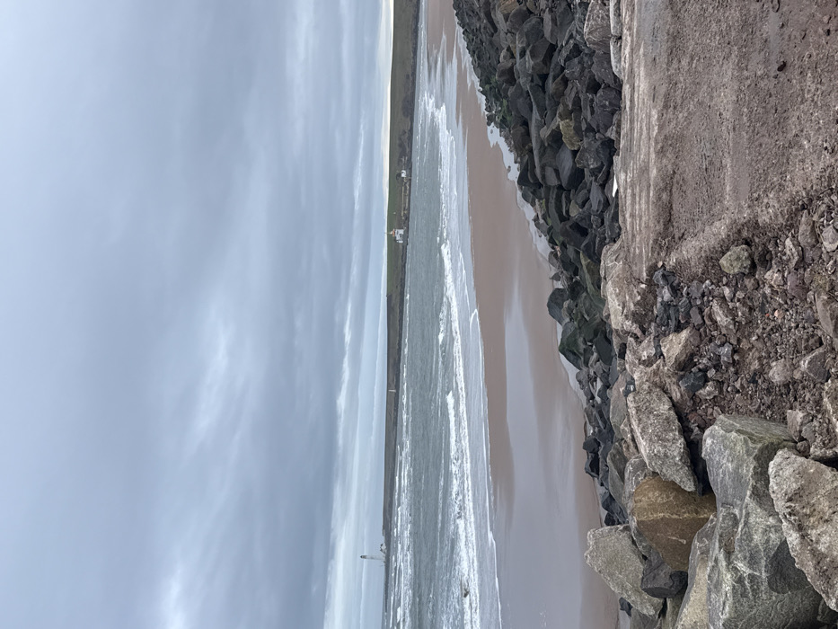 The image displays a coastal scene at Montrose, United Kingdom, specifically looking across Montrose Bay. The sky is entirely overcast with dull grey clouds. In the foreground, large, irregular dark grey and brown rocks, consistent with riprap, dominate the lower left and center, bordering a dark, rough ground surface that occupies the lower right. The midground features choppy, dark grey-green seawater with white breaking waves rolling onto a wide, crescent-shaped sandy beach, which appears dark and wet near the water's edge and lighter further inland. In the distance on the far left, a tall, slender white lighthouse is visible on a low headland. To the right, across the bay, another, shorter white lighthouse with a yellow cap, accompanied by smaller white buildings, stands on a grassy headland, from which a low sea barrier extends into the water. No individuals, vehicles, or specific wildlife are discernible within the scene.