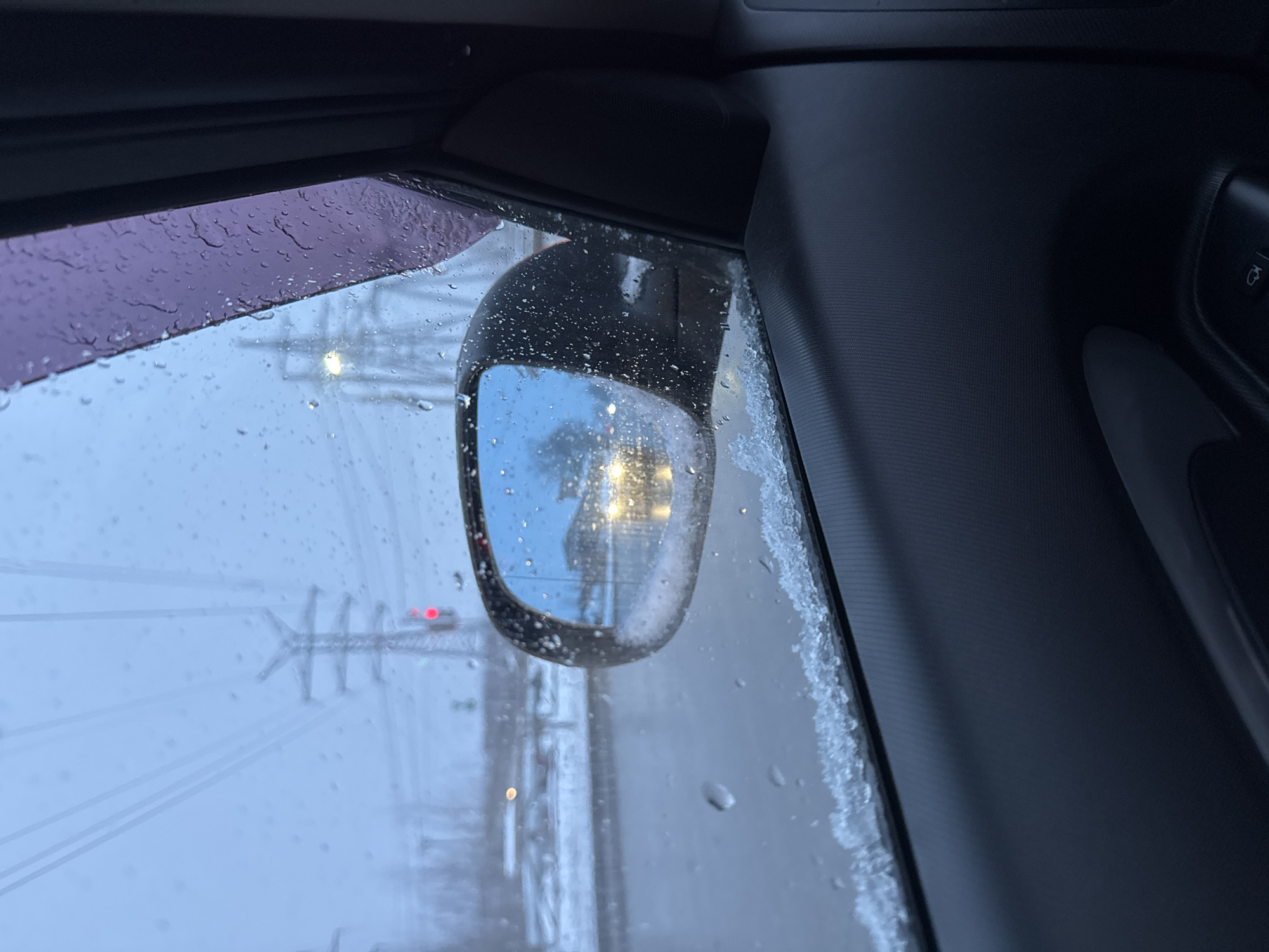 The photograph shows a view from inside a car on a snowy or icy day. Rain or snow is visible on the windows. The side mirror is covered in moisture and reflects street lights and infrastructure. A power line structure is visible through the driver's side window. The location is in Chaikovsky, Russia, based on the infrastructure and weather conditions. The image captures the bleak, wintery atmosphere from the perspective of a vehicle occupant. The focus is on the car’s side view mirror and the view through the window, with the car's interior partially visible.