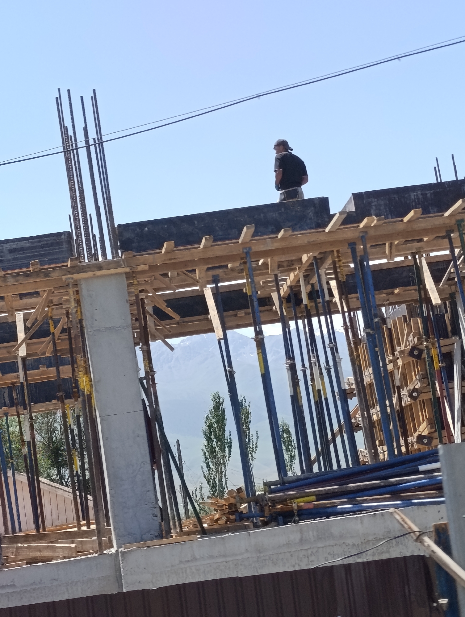 A man wearing a black shirt and a baseball cap stands on the second level of a construction site in At-Bashy, Kyrgyzstan. He is facing away from the camera. The construction site features concrete pillars and numerous wooden planks and metal poles used for support. In the background, mountains and trees are visible under a clear blue sky. The scene depicts ongoing construction work.