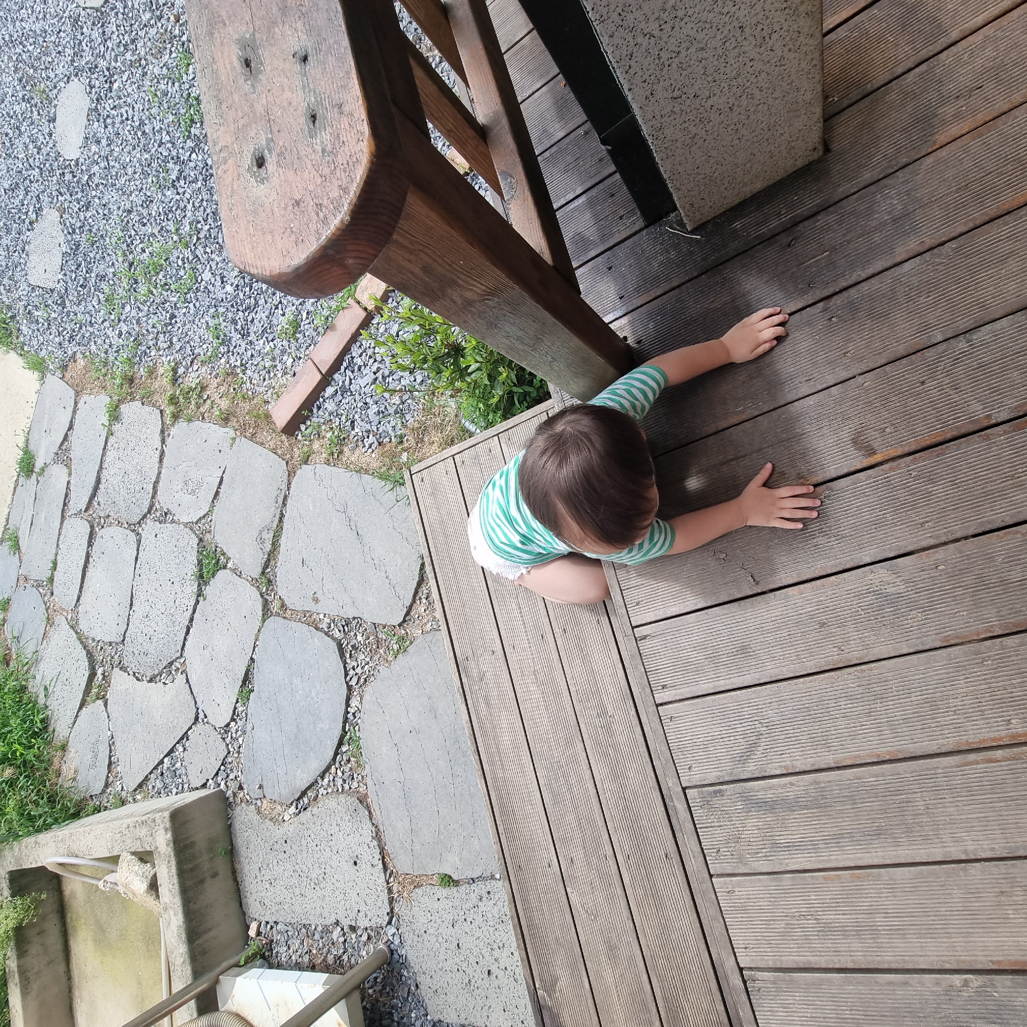 A toddler, wearing a green and white striped shirt and a diaper, is on hands and knees on a wooden deck. The toddler is facing away from the camera, looking down at the deck. The deck is made of weathered wooden planks. To the left of the deck, there is a pathway made of large, flat stones interspersed with gravel and grass. Beyond the stone pathway, there is a concrete trough with a hose. To the right of the deck, a portion of a wooden bench is visible, and further right, there is gravel and green foliage. The scene appears to be outdoors, possibly in a garden or outdoor seating area. The location is Dunnae-myeon, South Korea.