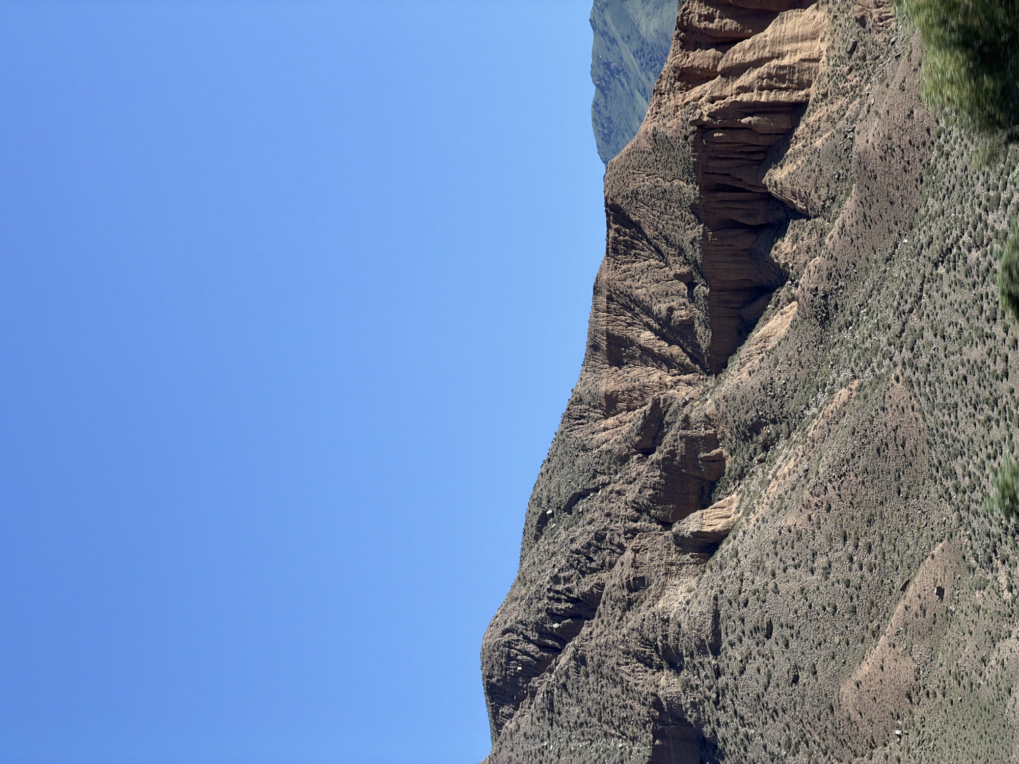 This is a landscape photograph of a mountainous region under a clear blue sky. The foreground features a rugged, reddish-brown mountain slope sparsely covered with dry vegetation. The mountain displays intricate geological formations, including vertical striations and eroded cliff faces, suggesting a history of weathering and erosion. In the background, further mountain ranges with a greener hue are visible, indicating a larger, natural environment. The image appears to be taken during daylight, with the sun casting shadows that highlight the textures of the terrain. The scene is devoid of human presence or any man-made structures, emphasizing the wild and natural character of the location. This geographical area is identified as being in the vicinity of Kok Moynok Eki, Kyrgyzstan.