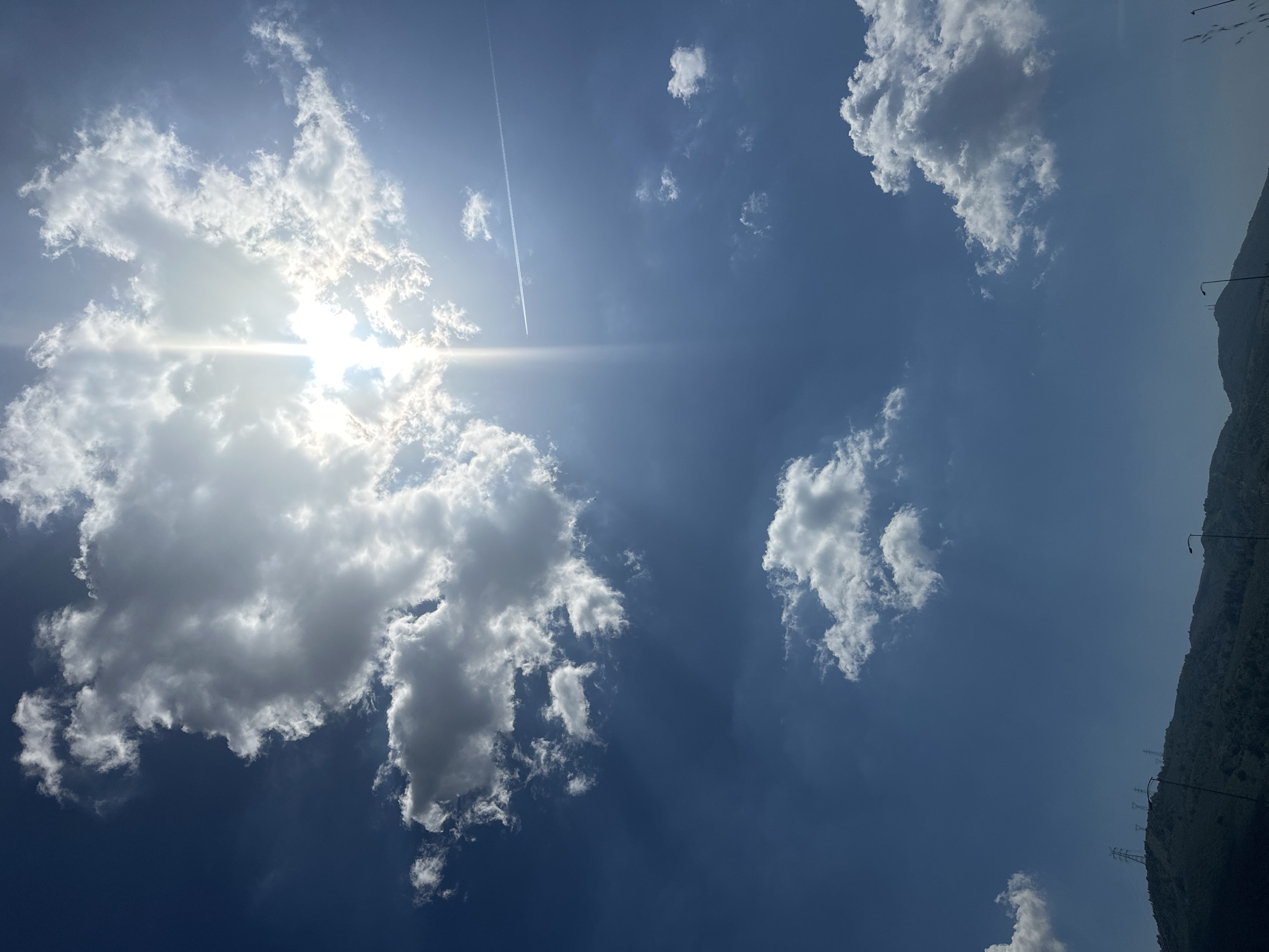 The image shows a daytime sky above Orhangazi, Turkey, with a bright sun obscured by white clouds. A contrail stretches across the upper left quadrant. To the right, the silhouette of a mountain range is visible, with power lines and street lights dotting the ridge. The sky is predominantly blue.