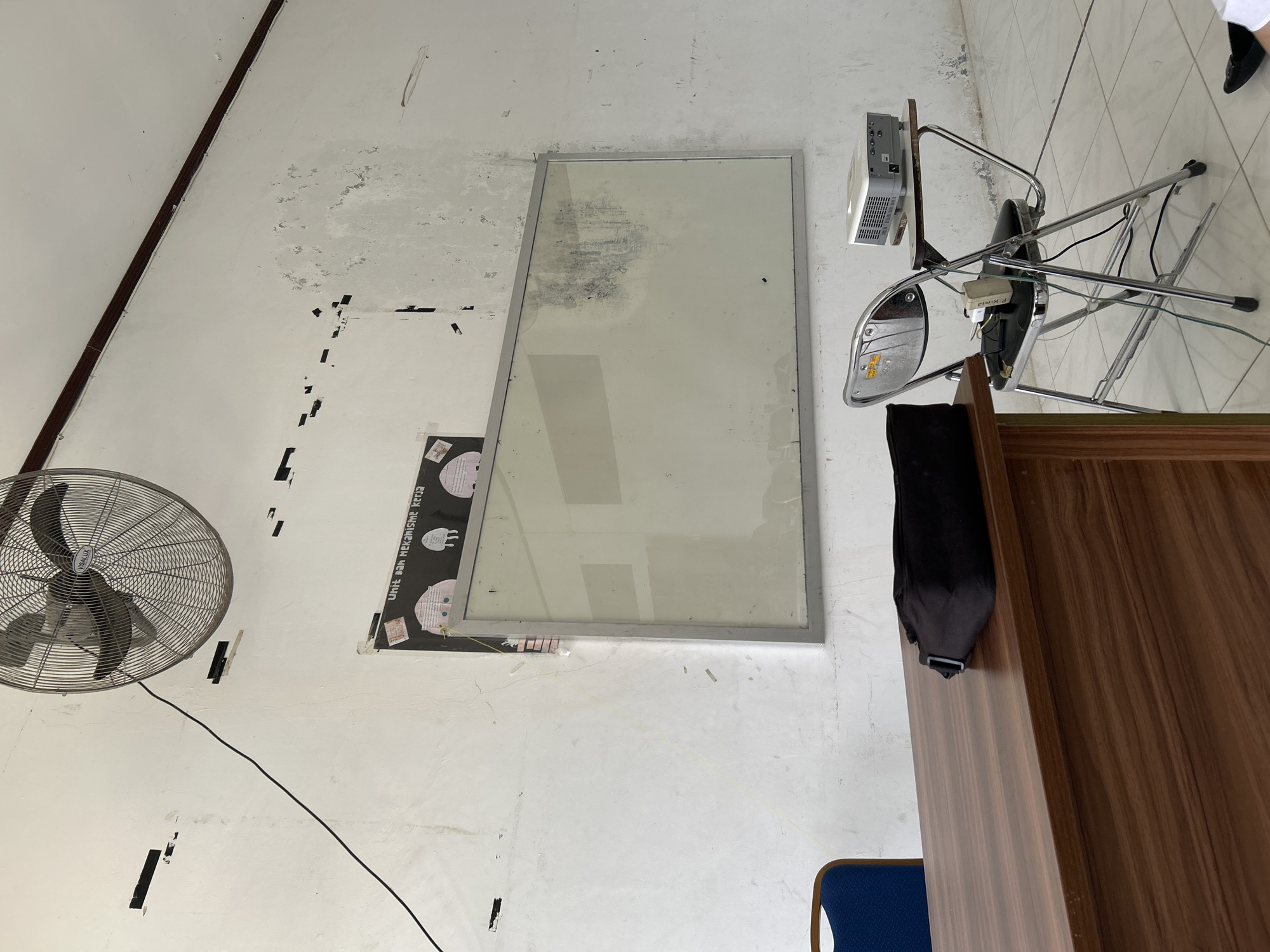 An indoor scene, likely a classroom or meeting room, is depicted with a worn white wall forming the primary background. The wall shows signs of peeling paint and discoloration, with several small black rectangular adhesive strips or holes scattered across it. A dark brown structural beam or trim runs diagonally along the upper left corner of the wall.
In the lower-left corner of the room, a large, grey metallic wall-mounted fan with the partial brand name "VISALUX" is visible. A black electrical cable extends from the fan along the wall.
Towards the center of the image, a large rectangular framed panel with a silver frame is mounted on the wall. Its surface, possibly a whiteboard or a reflective board, is heavily stained and smudged, particularly in its upper-left section. Below this panel, a portion of a black poster or chart is visible, displaying white text that reads "UNIT DAN MEKANISME KERJA" (Unit and Work Mechanism) and some abstract shapes.
On the right side of the room, an improvised setup features a white multimedia projector placed on a small wooden shelf. This shelf is precariously balanced on an overturned metal-framed chair with chrome legs and a dark seat. A green cable and partial text "P.K.N.G" are visible on the underside of the chair.
A large wooden desk or table with a prominent wood grain finish occupies the lower-right foreground. A small section of a blue-seated chair is visible beside it. In the far upper-right corner, a person's feet, wearing black shoes and white socks, are partially visible, suggesting human presence in the room.
The overall impression is one of a functional but somewhat dated or neglected interior space.