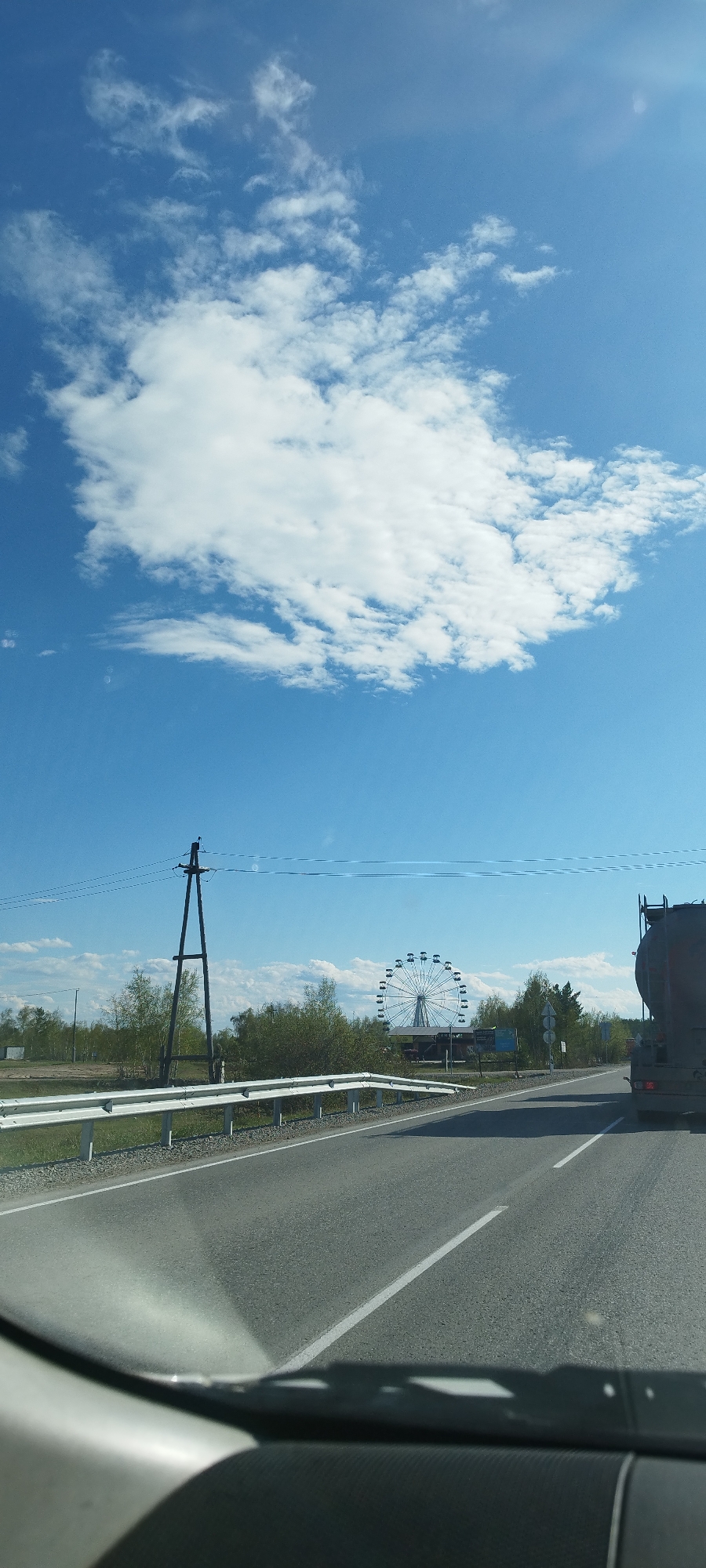 The image shows a road scene in Tekhtyur, Russia, on a clear, sunny day with scattered clouds in a blue sky. The view is from inside a vehicle, looking out onto a straight asphalt road with a solid white dividing line. A metal guardrail runs along the left side of the road. In the distance, a large Ferris wheel is visible behind trees and a utility pole. A tank truck is partially visible on the right side of the road, indicating traffic. The overall environment is rural or suburban, with trees and open fields in the background. No people are directly visible in the frame.