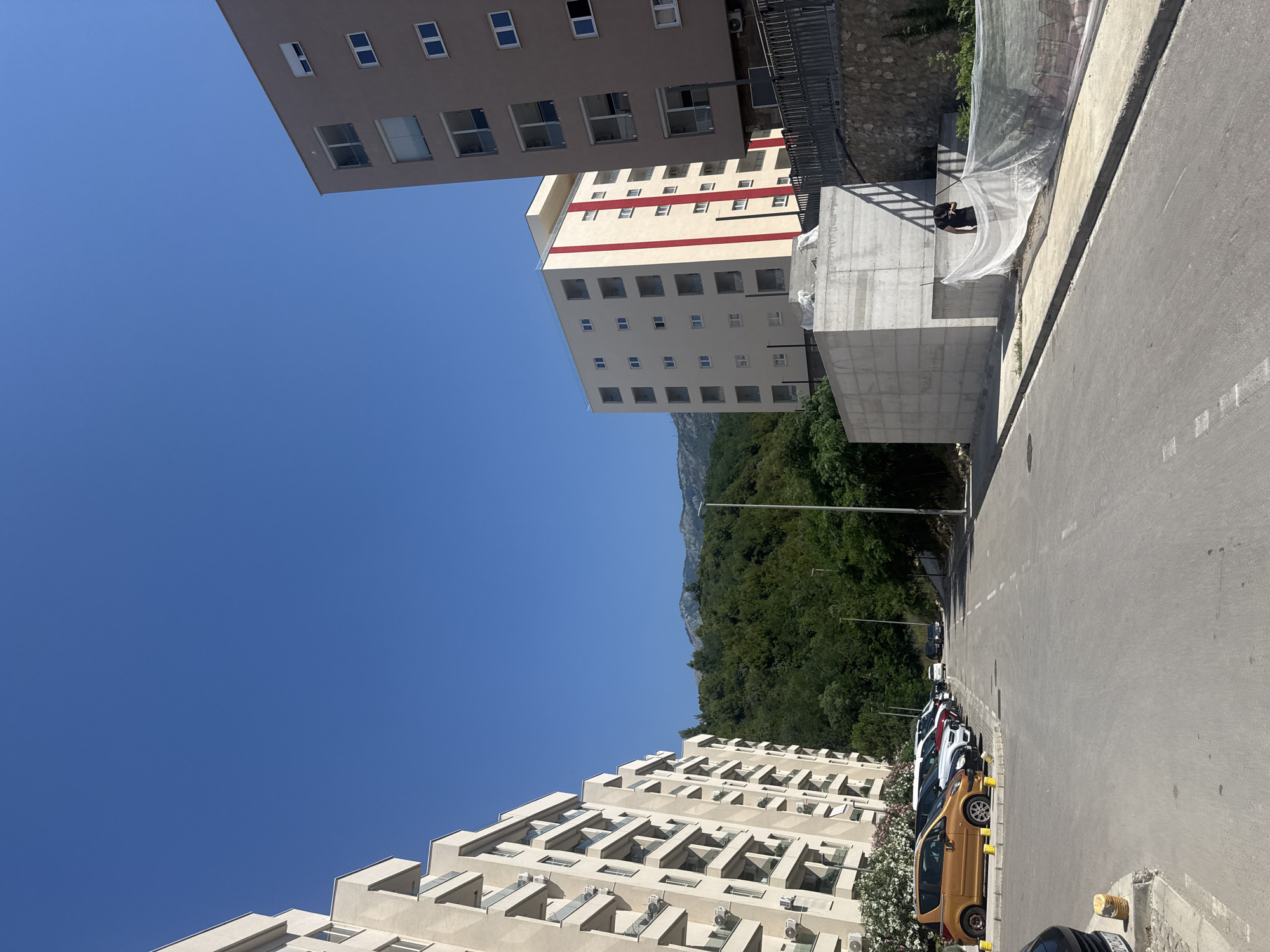 The image shows a street scene in Boreti, Montenegro, with several buildings and parked cars under a clear blue sky. The buildings are modern, with multiple stories and balconies. One building in the background has a striking red stripe running horizontally across its facade. A person is visible near a construction site in the middle ground, partially covered by netting. The street is paved, and several cars are parked along the side. The presence of air conditioning units on the buildings and lush green trees suggest a warm climate. The scene appears to be captured during daylight hours, likely in the late morning or afternoon, given the bright sunlight and shadows.