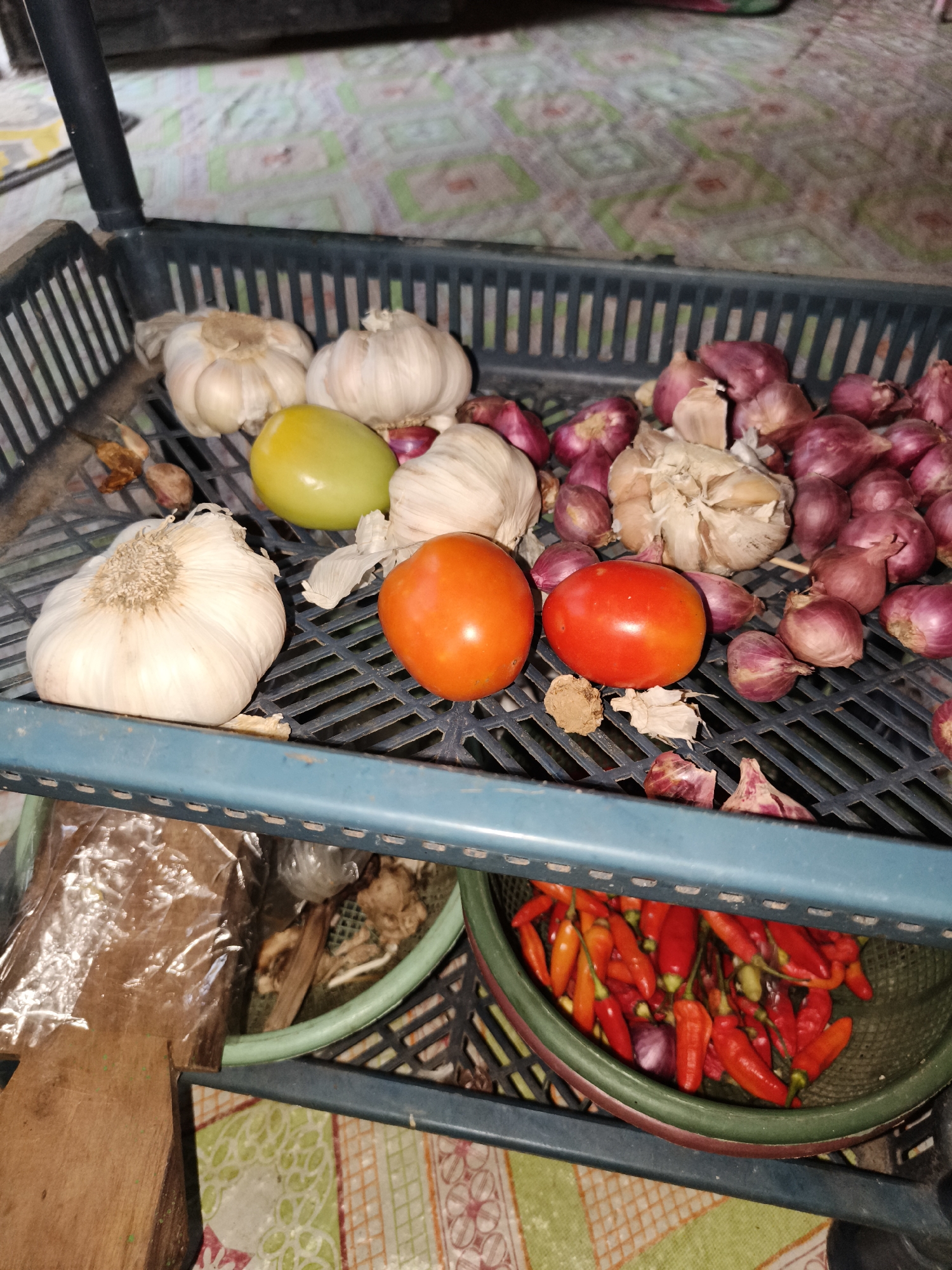 In Pelanjau Jaya, Indonesia, a close-up, eye-level shot displays a collection of fresh produce arranged in a black plastic two-tiered rack. The top tier holds several heads of garlic, a few red tomatoes, one greenish-yellow tomato, and a pile of small red onions. Some dried chili peppers and what appears to be ginger or galangal are also visible on this shelf. The bottom tier of the rack contains a green basket filled with numerous small red chili peppers. A wooden cutting board and some plastic-wrapped items are partially visible on the left side of the bottom tier. The background consists of a patterned mat.