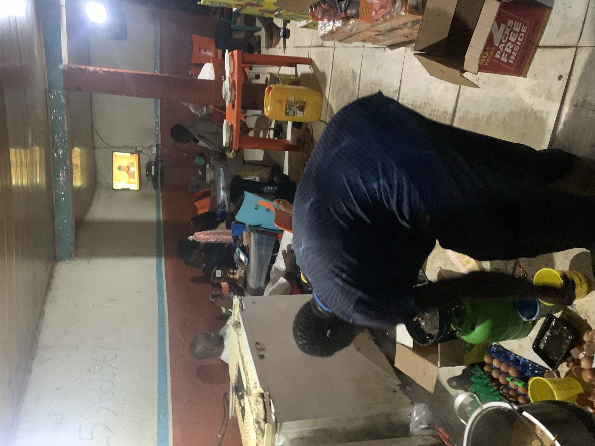 The image captures an indoor scene, likely an eatery or communal gathering space, illuminated primarily by artificial light, suggesting it is either evening, night, or a dimly lit interior. The environment appears functional and lived-in, characterized by simple furnishings and practical arrangements for food preparation and dining.
In the immediate foreground, a person clad in dark blue attire is bending over, actively engaged at what appears to be a food preparation or serving station. This area is quite cluttered, featuring a green gas cylinder, yellow buckets, and multiple trays containing eggs, with some broken eggshells visible, indicating ongoing activity.
Further into the background, several individuals, mostly men or boys, are seated at simple orange tables. White plates are visible on one of these tables, alongside a large yellow jerrycan. Many are oriented towards a small television screen mounted on a distant wall, which displays a close-up of a male figure, possibly a news anchor or public personality.
The floor is tiled, and an open cardboard box rests on it, clearly showing the text "40+ PACKS" and "PACKS FREE INSIDE." Along the left wall and extending onto the floor, some faded, handwritten numbers or letters are faintly discernible, resembling "530067." A white or metallic counter or fridge unit is also present on the left side of the frame. The artificial lighting creates strong contrasts and highlights across the scene.