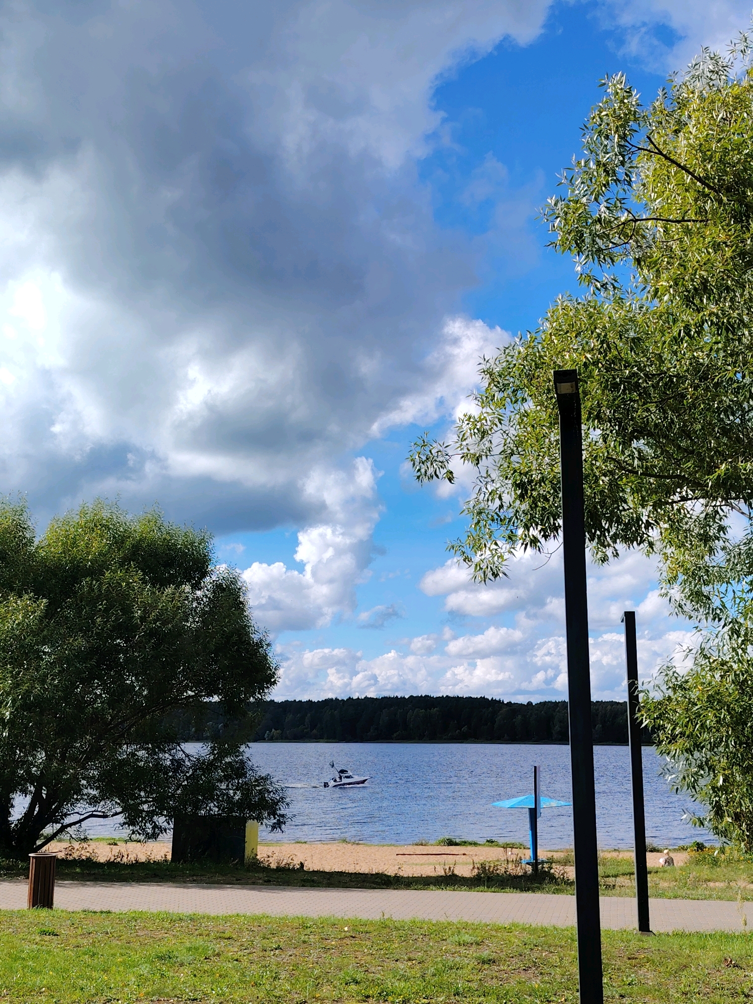 The image shows a serene waterscape with a calm lake or river, reflecting a partly cloudy blue sky. A small white boat with an outboard motor is moving across the water from left to right. The shoreline is sandy, backed by a dense forest of dark green trees. In the foreground, a paved walkway runs alongside a grassy area. Two dark, modern lampposts stand tall, partially obscuring a large, leafy green tree on the right side of the frame. Another large tree with green foliage is visible on the left, closer to the shoreline. A blue umbrella-like structure is present on the beach. The overall scene suggests a recreational area, likely a park or beach, situated by the water in the city of Konakovo, Russia, given the context.