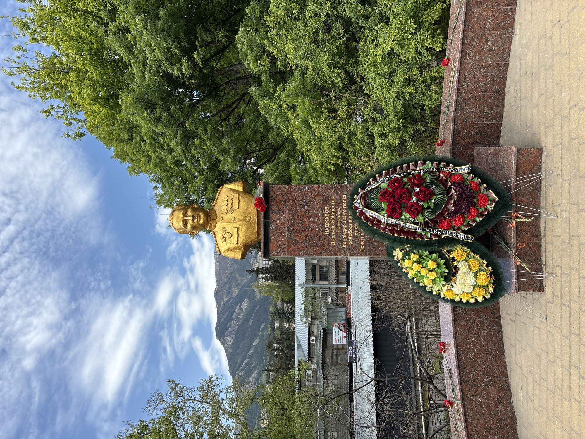The image shows a bust of a man in a military uniform, presumably a monument in the city of город Ялта, Ukraine. The bust is gold-colored and stands atop a red marble pedestal. Inscribed on the pedestal is text written in Russian. Two wreaths lie at the base of the monument, one with red flowers and another with yellow and white flowers. Behind the monument, a lush green tree fills the background. In the distance, a mountain range can be seen under a blue sky with scattered clouds. In the foreground, a sidewalk made of light brown bricks is visible. A building with several signs is visible in the background.