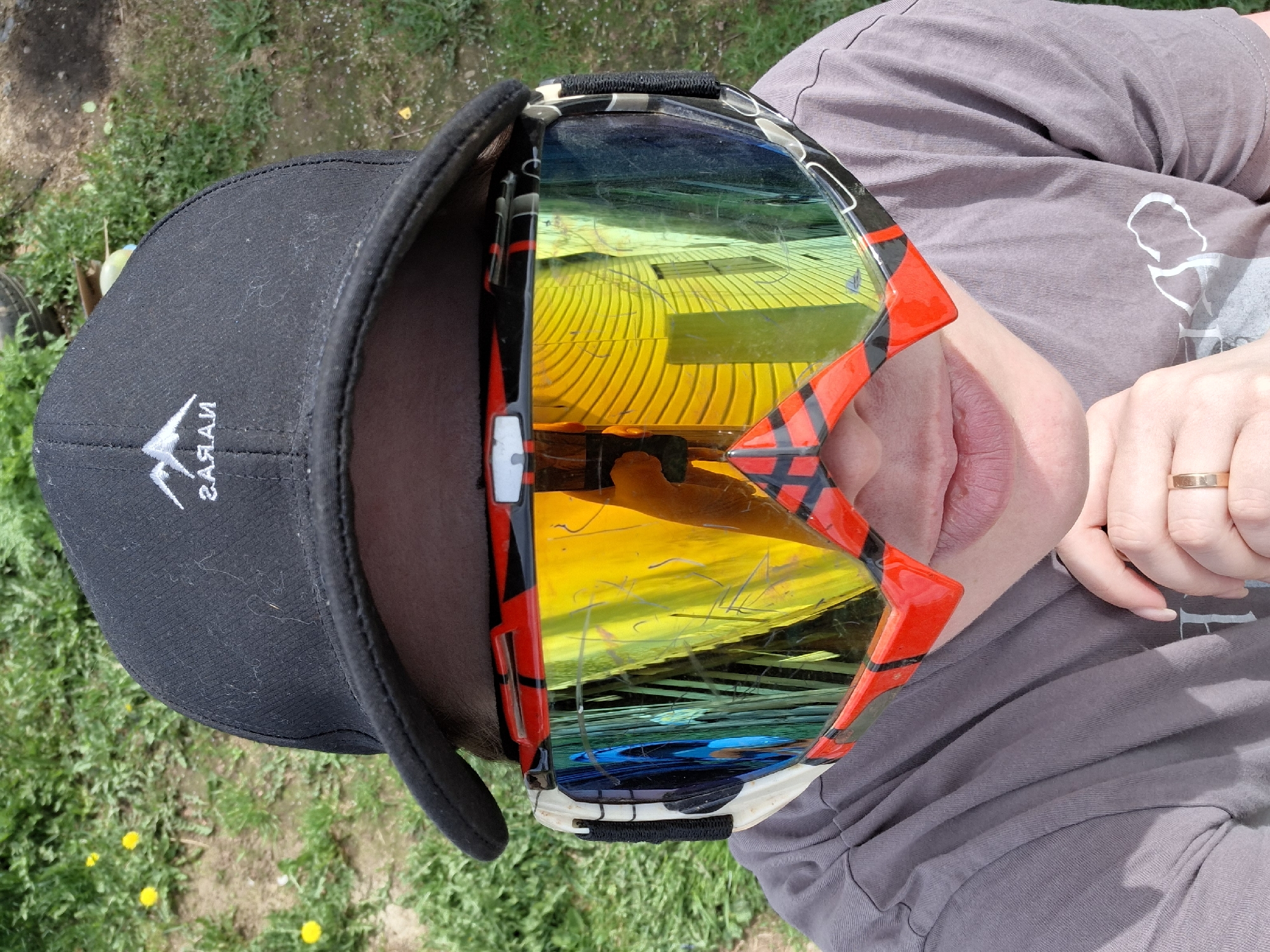 A person wearing a black baseball cap with white logo, a gray shirt, and red and black snow goggles with mirrored lenses is seen outdoors on green grass. The location and city are not discernible from the image.