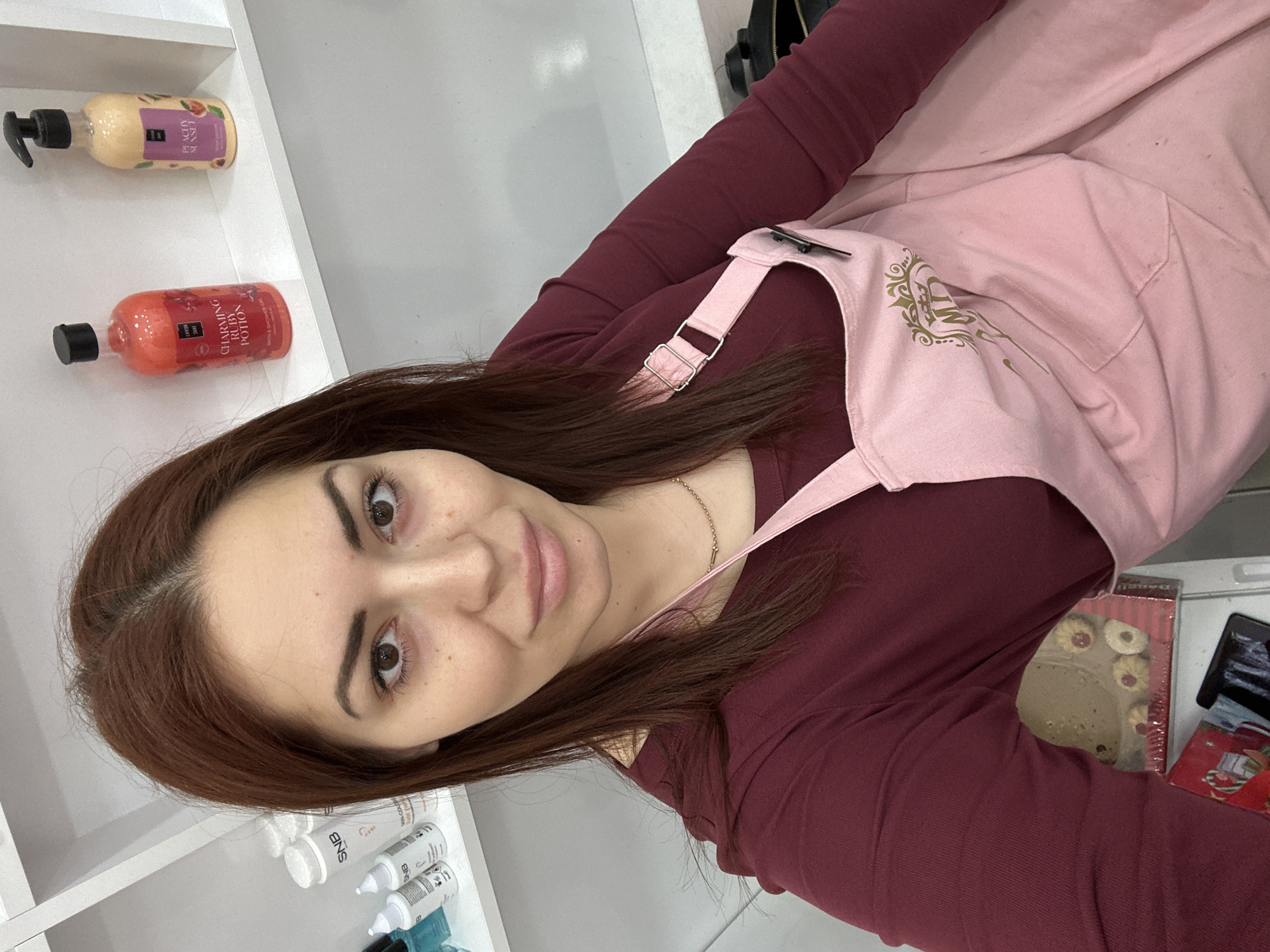 A woman is depicted wearing a burgundy, long-sleeved shirt and a light pink apron with a gold design. She appears to be in a commercial setting or personal care space, as evidenced by the presence of product bottles on a white shelf behind her and a package of cookies nearby. The bottles include products labeled "PEACHY SUNSET" and "CHARMING RUBY POTION". Additional small bottles with labels marked "SNB" are also on the shelf. The location is not evident from the image alone.