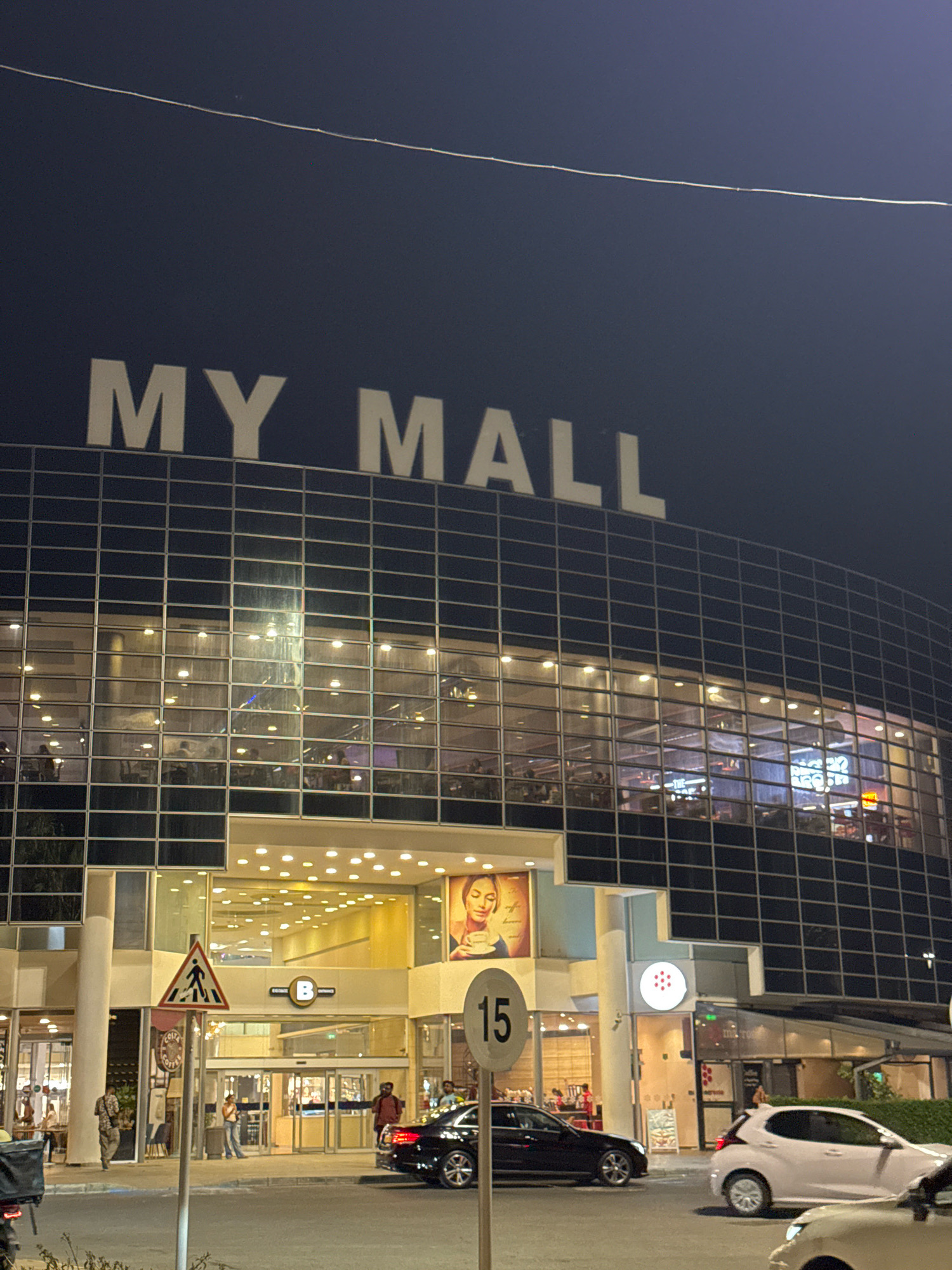 The image depicts the exterior of the MY MALL in Limassol, Cyprus, at night. The large, modern building features a dark facade with a grid pattern of windows. The prominent "MY MALL" sign is illuminated in white lettering against the dark background. The windows of the mall reveal illuminated interiors with visible lights and silhouettes of people, suggesting it is open for business.
In the foreground, a street scene is visible with a few cars parked or driving. A black sedan is positioned in the middle ground, and a white hatchback is to its right. A white sign with the number "15" is mounted on a pole in the foreground, likely indicating a speed limit or parking restriction. Another traffic sign with a pedestrian crossing symbol is also visible.
The entrance to the mall is a large, glass-enclosed area with automatic doors. People are seen entering and exiting the building. A sign with the letter "B" in a circle is displayed above the entrance. Adjacent to the entrance are storefronts, one of which appears to be a "Costa Coffee." A large poster featuring a woman holding a coffee cup is displayed on the exterior wall of the mall near the entrance. The overall scene suggests a busy commercial area at evening or night.