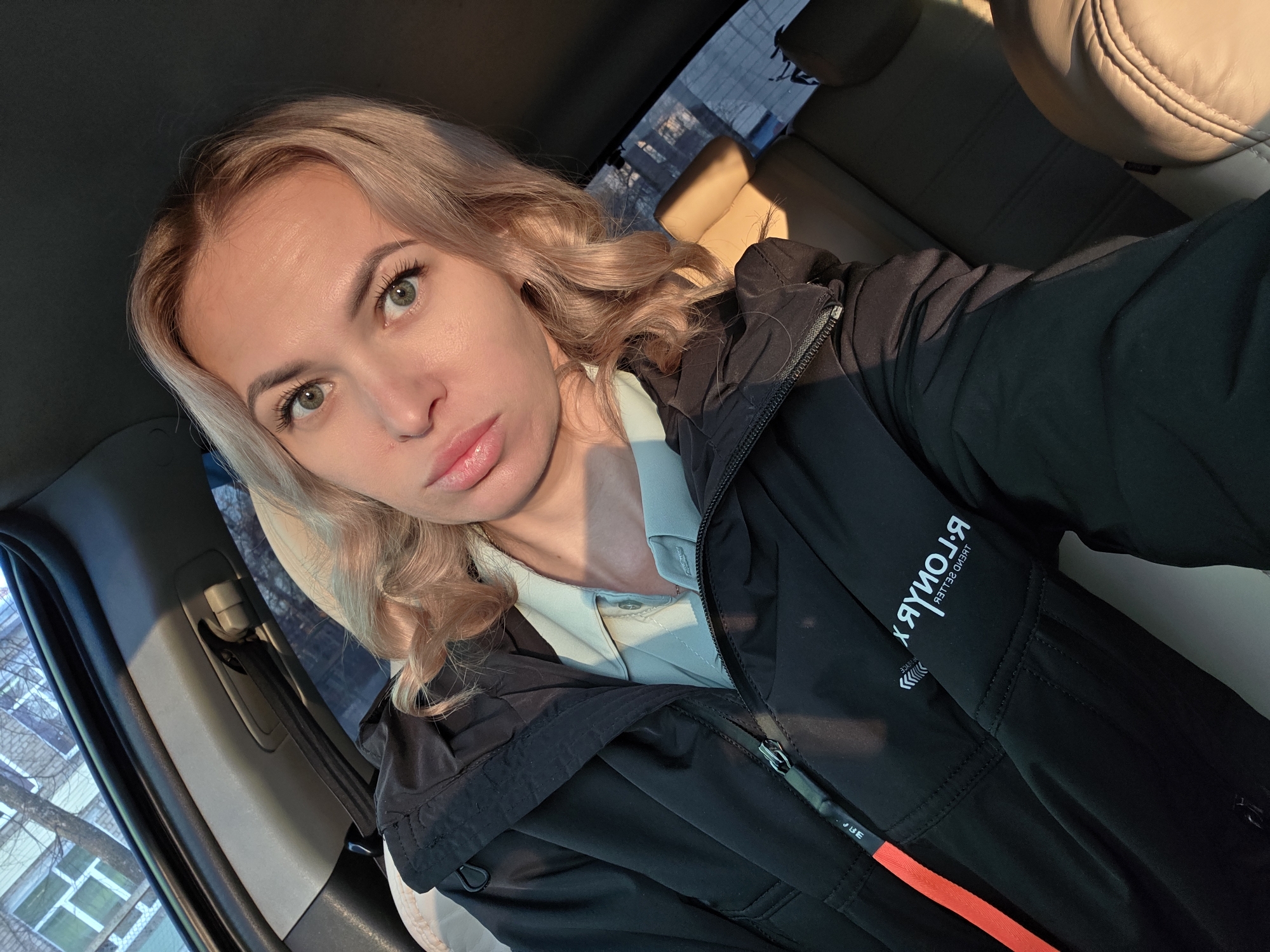 A young adult female with blonde, wavy hair and green eyes is seated in the front passenger seat of a vehicle in Chaikovsky, Russia. She is wearing a light green collared shirt under a black hooded jacket. The jacket features an orange vertical stripe on the wearer's left side and white, mirrored text, "R. LONOR" and "TREND SELLER", on the wearer's right sleeve. She looks directly at the camera with a slightly pursed expression. The vehicle's interior has light-colored upholstery, and a seatbelt mechanism is visible in the upper left. Strong sunlight illuminates the left side of her face. Through the vehicle's windows, a multi-story building with numerous square windows and a tree with bare branches are visible, indicating an urban environment.