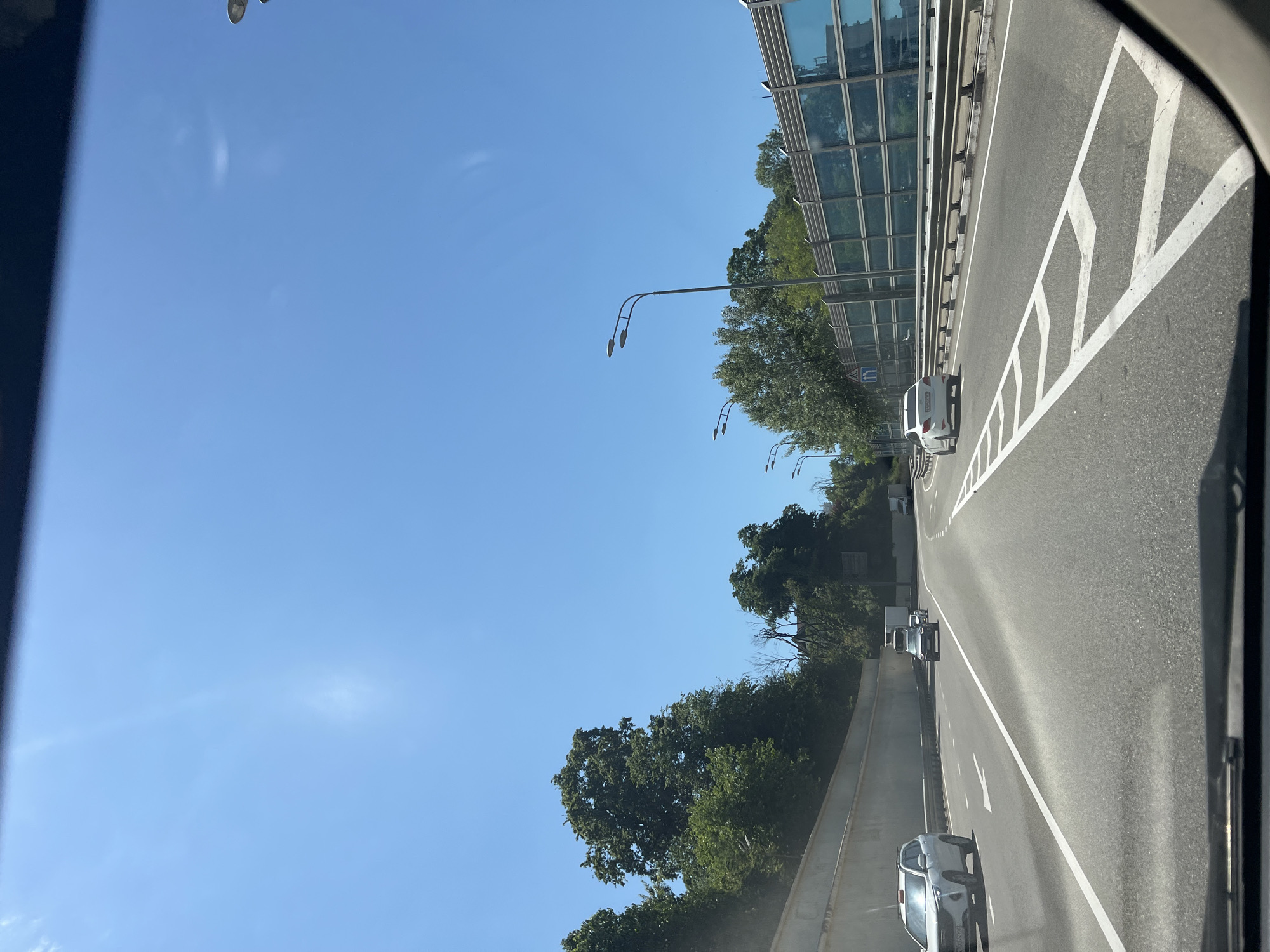 The image depicts a bright, sunny daytime scene from the perspective of a vehicle traveling on a multi-lane highway. The sky, occupying the upper portion, is clear and a vibrant blue with only a few faint, wispy clouds.
On the left side of the highway, a tall, modern structure with numerous rectangular glass panels, likely a sound barrier, runs parallel to the road. Behind this barrier, a dense line of green trees and bushes stretches into the distance. Several streetlights with dual lamp heads extend over the road from this side. A small blue road sign with white arrows, one pointing left and one upwards, is partially visible amongst the trees.
The asphalt road features clear white markings, including solid lines, dashed lines, and prominent chevron patterns on what appears to be the rightmost lane or shoulder, possibly indicating a merge area or a restricted zone. Several cars are visible on the highway, with a white SUV or hatchback in the foreground on the right lane. Other cars, including another white vehicle and a silver/grey one, are further ahead on different lanes.
The overall scene suggests a pleasant day for travel on a well-maintained road system, possibly on the outskirts of an urban area, given the presence of the sound barrier and highway infrastructure. There are no people visible, and no readable text is discernible on any objects or vehicles.