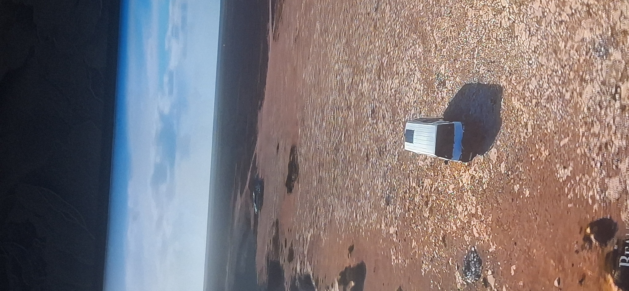 The image displays an aerial view of a desolate, arid landscape. The foreground is characterized by dry, cracked earth with scattered rocks and sparse vegetation, giving it a desert-like appearance. A white van with a blue front is positioned in the center of the frame, casting a dark shadow on the ground. In the background, the landscape transitions to darker, possibly more barren terrain, with a subtle gradient towards the horizon. The sky above is partly cloudy with patches of blue sky visible, suggesting daylight. There is no indication of the city of Bila Tserkva, Ukraine, or any urban elements within the scene.