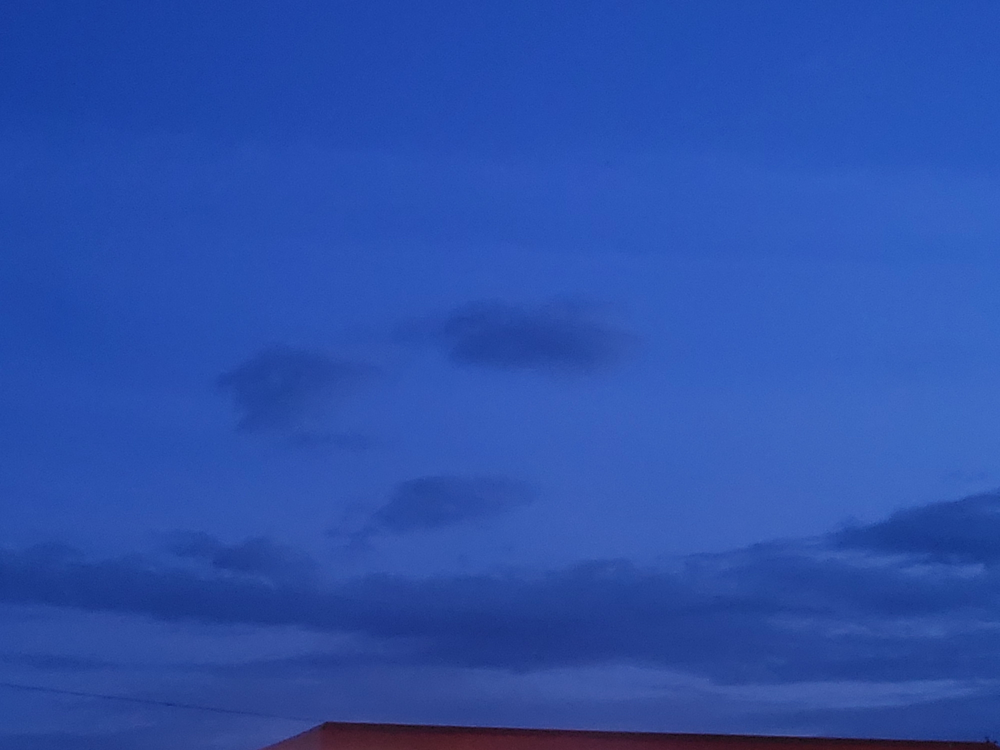 A nighttime photograph captures a deep blue sky with scattered dark clouds. In the lower portion of the frame, the edge of a building with a red-tiled roof is visible. The scene is devoid of any people or discernible activities, presenting a quiet and still environment. The location is identified as Jelgava, Latvia.