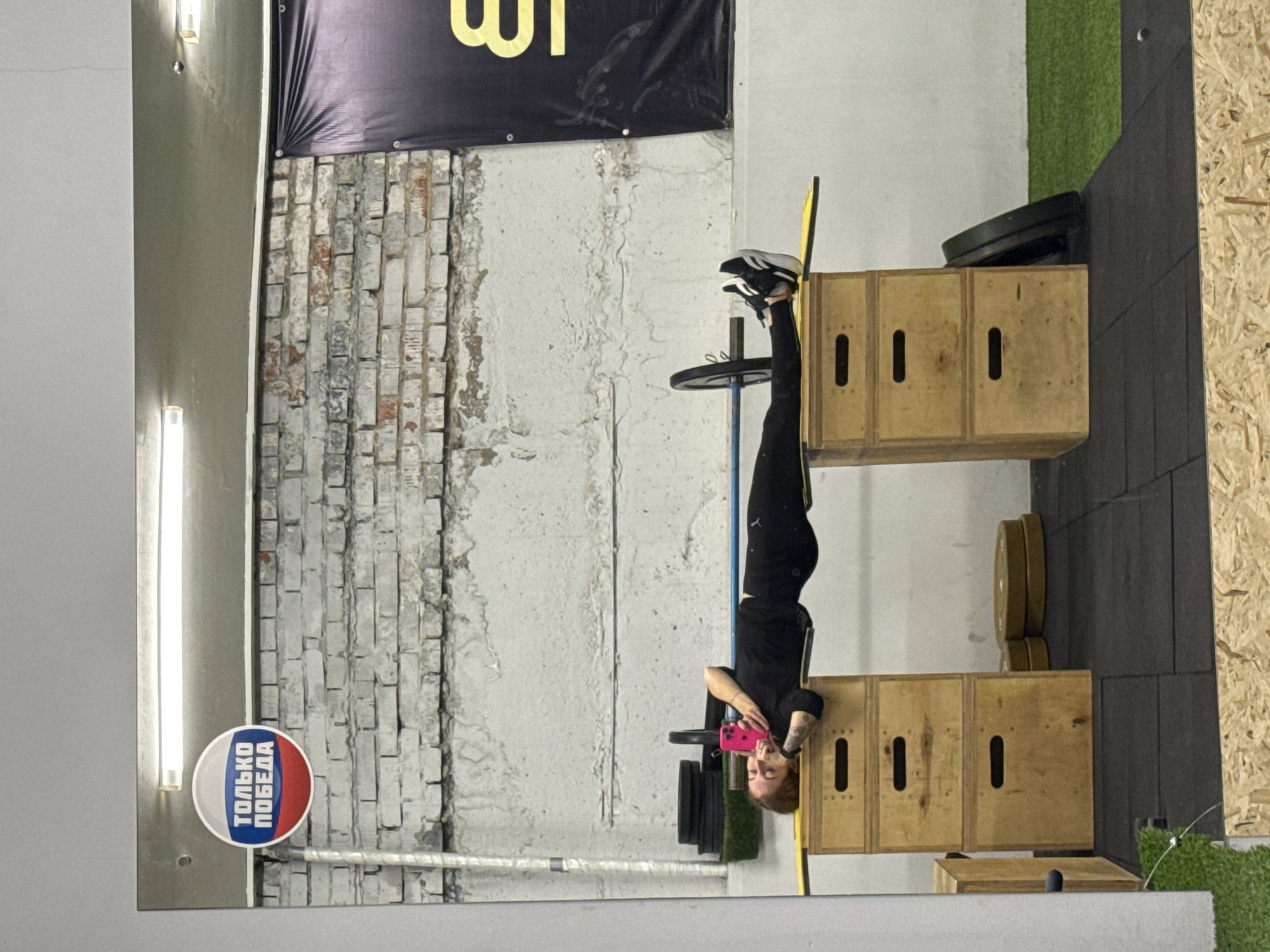 This media file depicts a gym or fitness studio in Chaikovsky, Russia, captured through what appears to be a large mirror, causing the main subject to be reflected upside down. The central figure is a person, likely a woman, dressed in black athletic attire and black and white sneakers. She is performing an inverted exercise, with her feet propped on a stack of wooden plyometric boxes. While in this challenging pose, she is holding a bright pink smartphone, suggesting she might be taking a selfie or recording her workout.
The setting showcases an industrial-style interior with an exposed brick wall, a painted white wall, and sections of green artificial turf and OSB paneling. Various gym equipment is visible, including dark and gold-colored weight plates stacked near the wooden boxes, and a yellow bar leaning against them. Overhead, a large black banner with partial yellow Russian text, "ШТ," is displayed against the brickwork. The space is brightly lit by fluorescent strip lights.
A prominent detail is a circular sticker on the wall, featuring horizontal white, blue, and red stripes, reminiscent of the Russian flag. The sticker bears the Russian text "ТОЛЬКО ПОБЕДА," which translates to "ONLY VICTORY," reinforcing the local context of Chaikovsky, Russia. The overall scene conveys an atmosphere of fitness and determination.