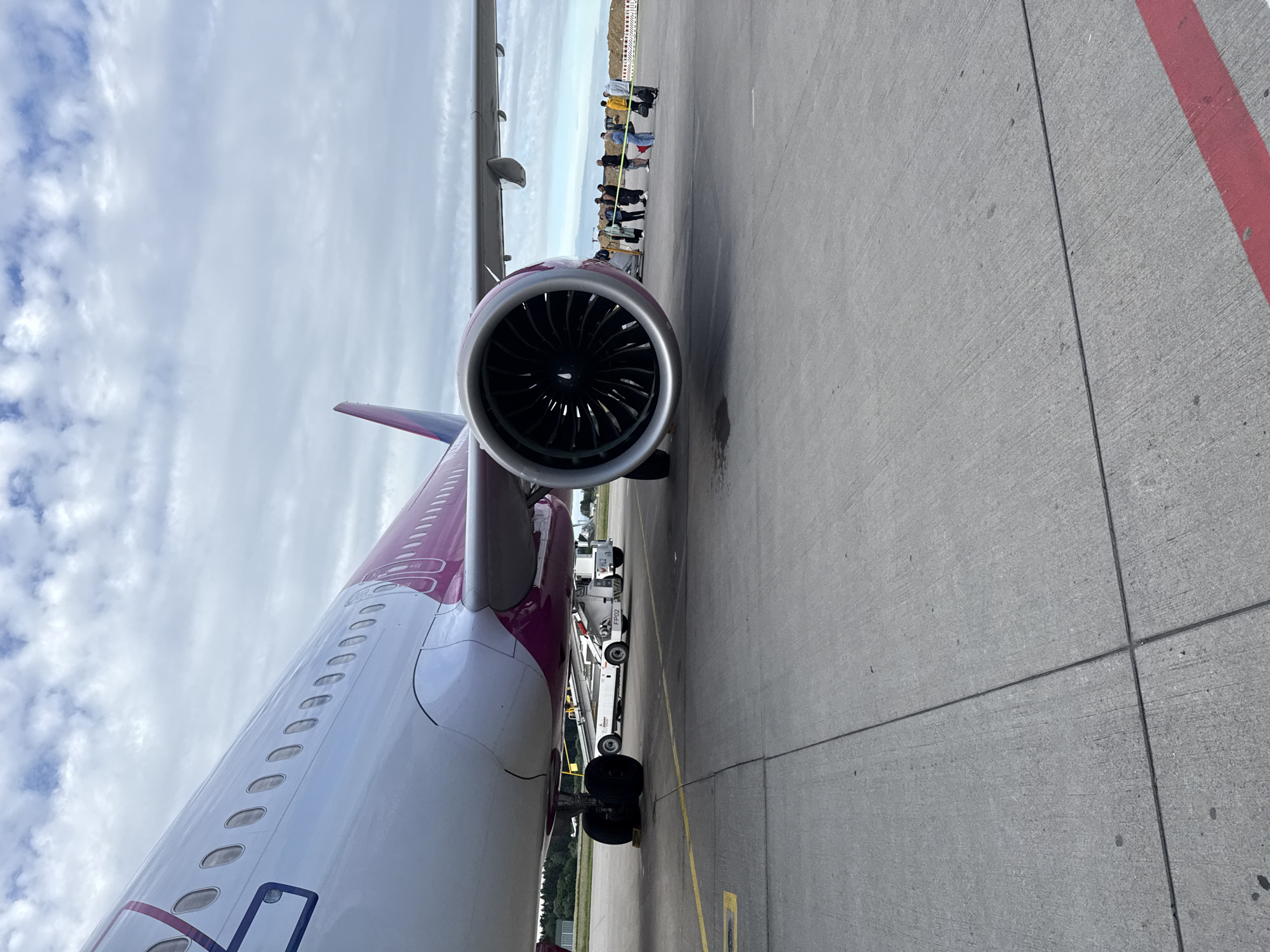 An airport tarmac at Künersberg, Germany, features a close-up view of the engine and fuselage of a pink and white passenger airplane. The airplane's landing gear is visible, as is a ground support vehicle and a boarding ramp. A group of passengers, some carrying luggage, stand near the aircraft, seemingly waiting to board. The sky above is filled with white, scattered clouds. The tarmac is concrete with yellow and red painted markings.