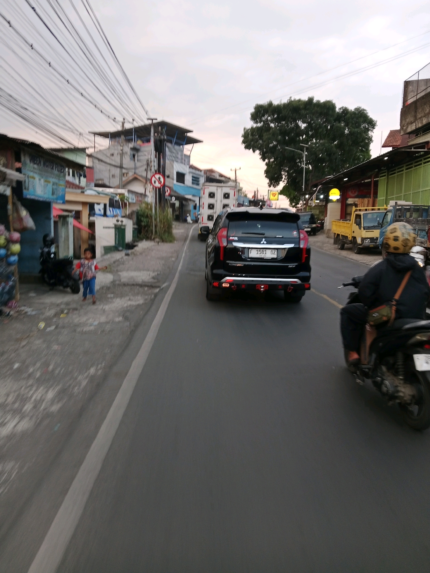 The image depicts a street scene in Sirnagalih, Indonesia. A black Mitsubishi Pajero Sport SUV with license plate F 1581 OZ is traveling on the right side of the road. In the foreground, a person on a motorcycle, wearing a dark jacket and a patterned helmet, is also traveling in the same direction. On the left side of the road, a young child wearing a patterned shirt and blue pants is standing on a sidewalk next to a motorcycle and a stall displaying colorful spherical items. Several buildings line the street, including one with a green facade and another with a tiled roof. Overhead, a complex network of electrical wires is visible against a cloudy sky. Yellow and blue trucks are parked on the right side of the road further ahead.