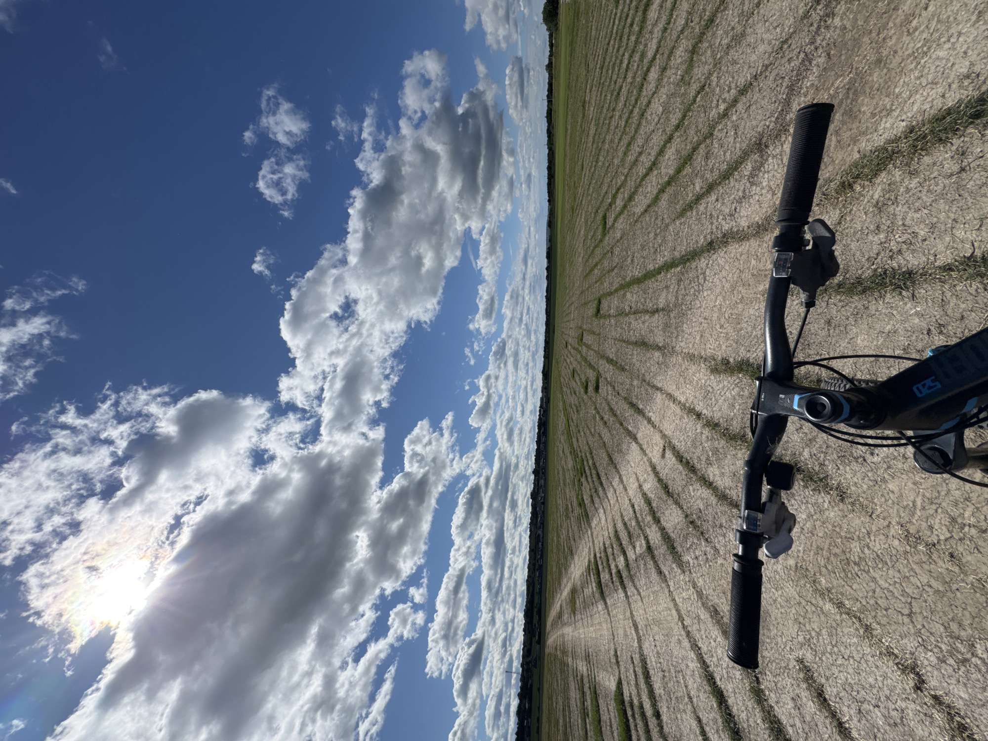 The image depicts a first-person perspective of someone riding a bicycle on a dirt path through a field. The field is lined with rows of young green plants. In the background, there is a distant treeline and buildings, suggesting a rural or semi-rural setting near a populated area. The sky is a vibrant blue, filled with scattered white and grey clouds. The sun is visible, partially obscured by clouds, casting bright light and lens flare. The handlebars and part of the bicycle frame are in the foreground, indicating the cyclist's point of view. The overall scene is outdoors, under a bright, cloudy sky.