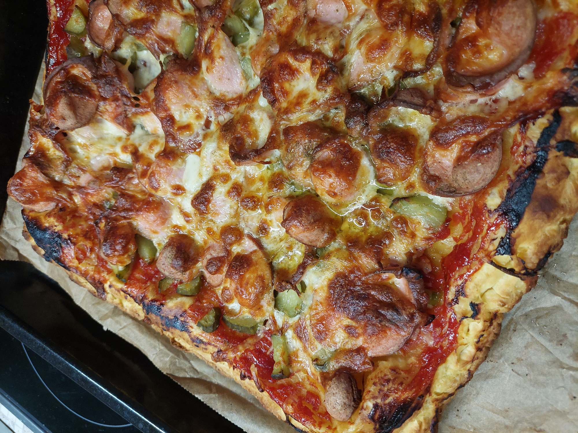 A close-up, high-angle shot depicts a pizza. The pizza appears to have a rectangular shape. It is topped with melted cheese, sliced sausage, and pieces of green pickle. The crust is golden brown and slightly charred in places. It is resting on a piece of parchment paper and a black baking tray. The image does not contain enough information to determine its location in Plovdiv, Bulgaria, nor any people, actions, or interactions.