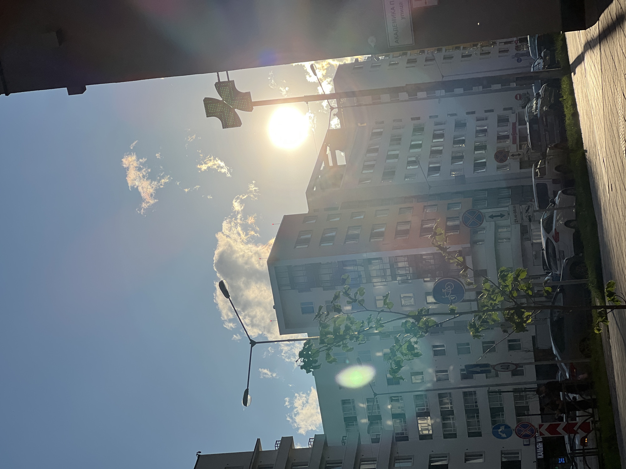 The image captures a sun-drenched urban scene in Yekaterinburg, Russia, likely during the daytime. The bright sun, positioned high in the clear blue sky with scattered white clouds, creates a strong lens flare and glares on the modern buildings. The perspective is from below, looking upwards, with a dark overhang in the foreground partially obscuring the view.
Dominating the frame are several multi-story buildings with numerous windows, reflecting the sunlight. One building appears to be under construction or renovation, with some scaffolding visible. A streetlamp with two lights stands tall, and a pedestrian crossing signal with green lights is also visible. Trees with sparse green foliage are interspersed within the scene, with one branch extending into the lower right portion of the frame, catching the sunlight.
The street level shows parked cars and a sidewalk made of paving stones. Several traffic signs are present, including a bicycle lane sign, a sign with a circle and diagonal line (prohibiting entry), and other road signs with text and symbols. The overall impression is one of a bright, active city day, with the sun being a prominent element influencing the lighting and atmosphere.
Visible text includes "АКАДЕМИКА ПА..." on a sign near the top of the frame and a sign with "21.00-00.00" possibly indicating a time restriction. The presence of Russian text and the location context confirm this as Yekaterinburg. The image conveys a sense of everyday urban life bathed in strong sunlight.