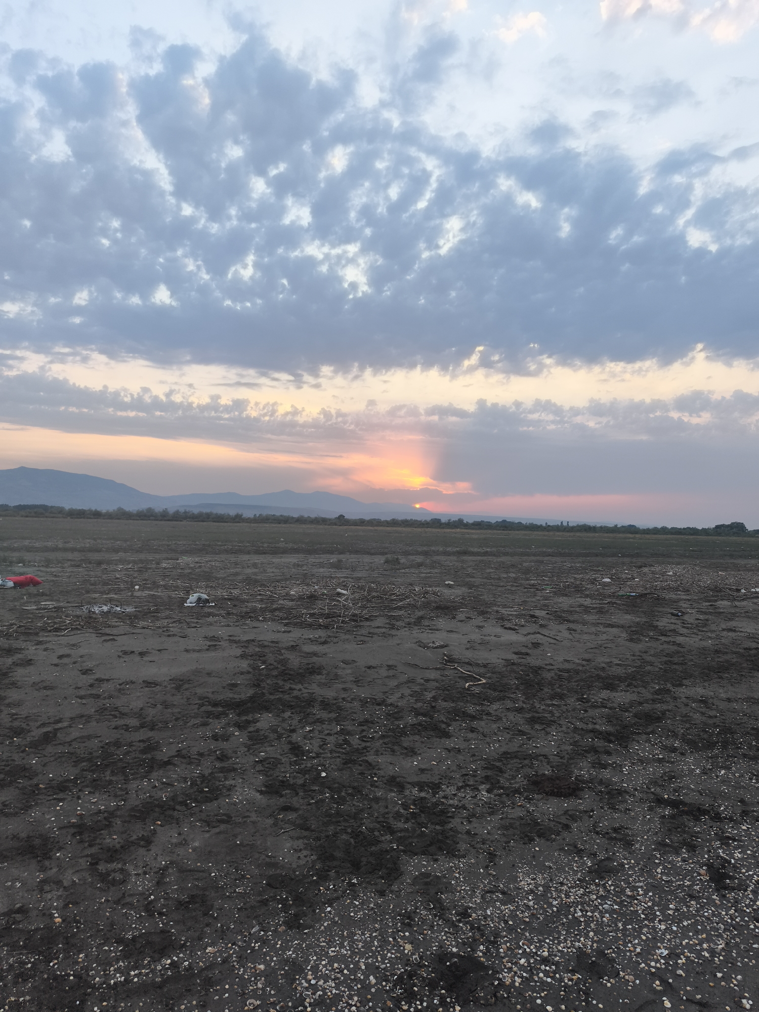 The image shows a desolate, dark, sandy expanse littered with debris, including plastic bags, branches, and what appear to be seashells. In the distance, rolling hills or mountains are visible under a dusky sky. The sky is partly cloudy, with the sun setting and casting a soft, orange and pink glow through a gap in the clouds. The overall impression is one of a neglected or undeveloped area on the outskirts of Balaca Həmyə, Azerbaijan, at dusk.