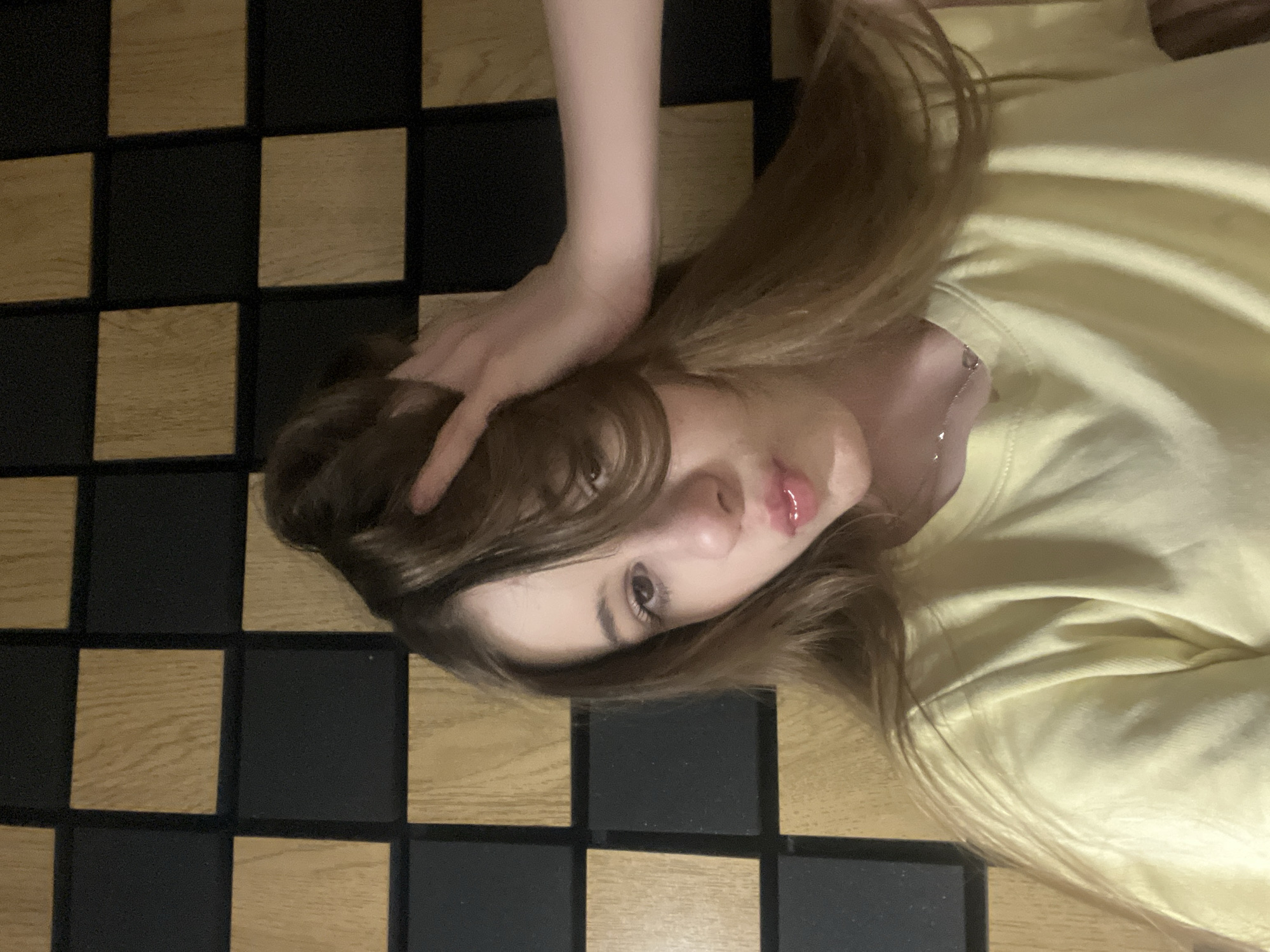 The image is a selfie of a young woman with light brown hair, wearing a yellow t-shirt and a silver necklace. She has her hand in her hair and is puckering her lips. She is standing in front of a wall with a checkered pattern of light brown and black squares. The background appears to be an indoor setting, possibly a room with acoustic paneling. There is no identifiable information or context that would allow for the determination of her location in the city of Dnipro, Ukraine.