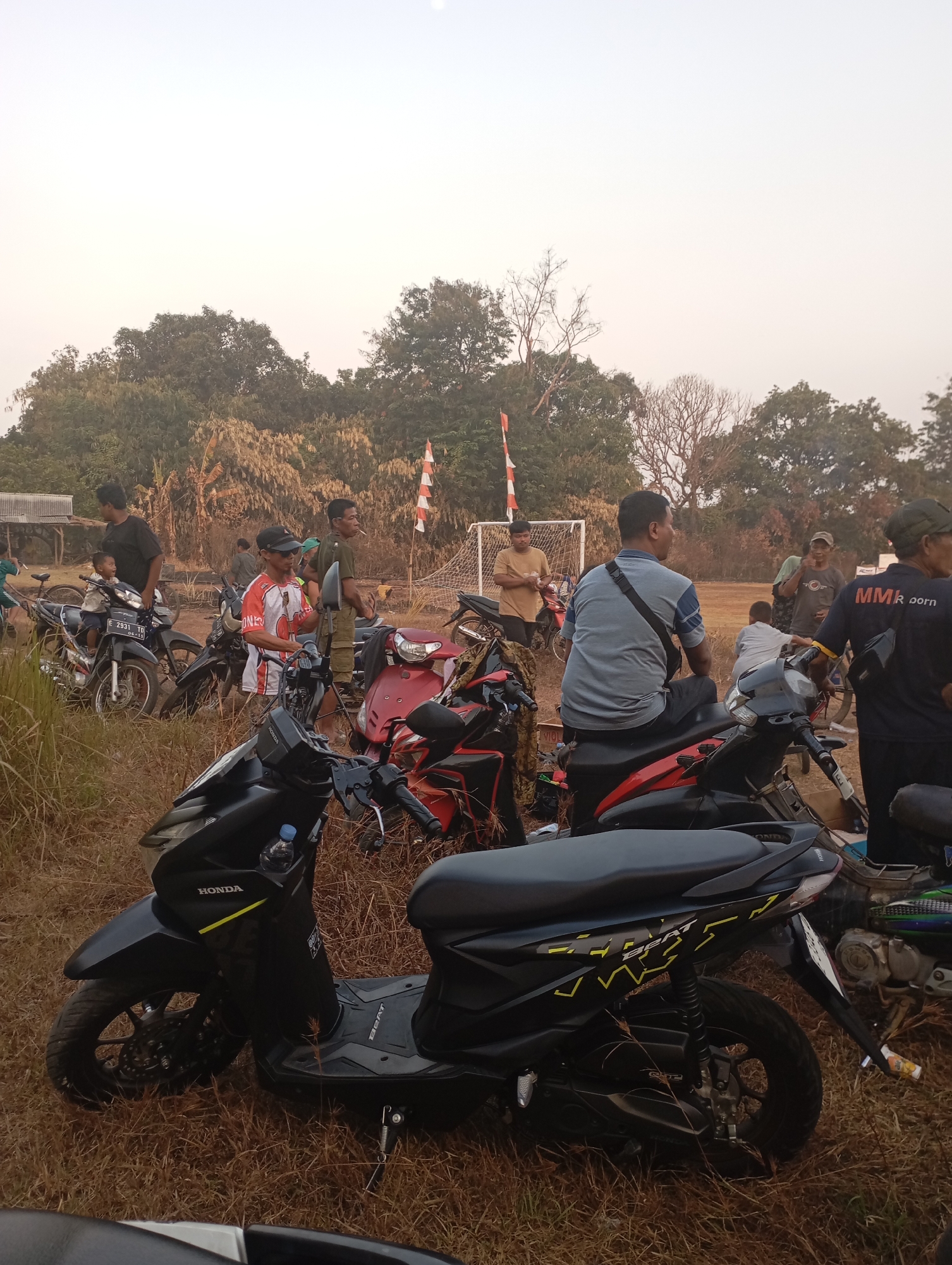 A group of people are gathered in a grassy, outdoor area, likely a field, with several motorcycles parked around them. The scene appears to be in a rural or semi-rural setting, possibly in Kecamatan Haurgeulis, Indonesia, given the context.
The subjects include several adult men and one child. Many of the men are standing or sitting on or near the motorcycles. One man in the foreground wears a grey shirt and black pants, with a black bag slung across his shoulder. Another man, partially visible in the lower right, wears a black shirt with "MMR porn" printed on the back. A man in the mid-ground wears a red and white jersey and a black cap. A young child is seated on a motorcycle in the left-middle of the image. In the background, more people are visible, and there are trees and foliage. A soccer goal and goal net are visible in the background, suggesting the area might be a sports field or a common gathering space.
The time of day appears to be late afternoon or early evening, with the sun low in the sky, casting a warm, diffused light. The sky is pale and hazy, suggesting possibly a dusty or humid atmosphere. The ground is covered in dry grass and some patches of bare earth.
The activity seems casual, with people interacting and observing their surroundings. There is no strong indication of a formal event, but rather a relaxed social gathering. The presence of motorcycles suggests transportation and possibly a shared interest in them.
Notable details include the distinct markings on the motorcycles, such as the "HONDA" logo and "BEAT" model name visible on the scooter in the foreground. The red and white striped flags in the background are also a visual element.
The overall mood seems calm and ordinary, with no overt signs of distress or excitement. The setting is natural and somewhat unkempt, reinforcing the rural context.