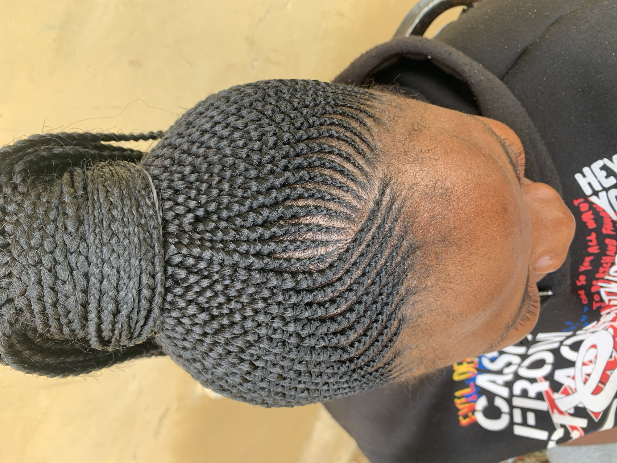 This media file shows a close-up, overhead view of a person's head, featuring an intricate braided hairstyle. The braids are small and neat, styled in a pattern that curves from the forehead towards the back of the head. A portion of the hair is gathered into a thick bun at the back, secured with what appears to be a braided band. The person is wearing a black hoodie with graffiti-style text in various colors, including white, red, and yellow. Some of the visible text reads "HEY...", "YOU", and "CAS...", with other words partially obscured. The background is a plain, light yellow surface, suggesting an outdoor setting. The lighting is bright, indicating daytime. The overall impression is of a well-maintained and fashionable hairstyle. The location context is Jos, Nigeria.