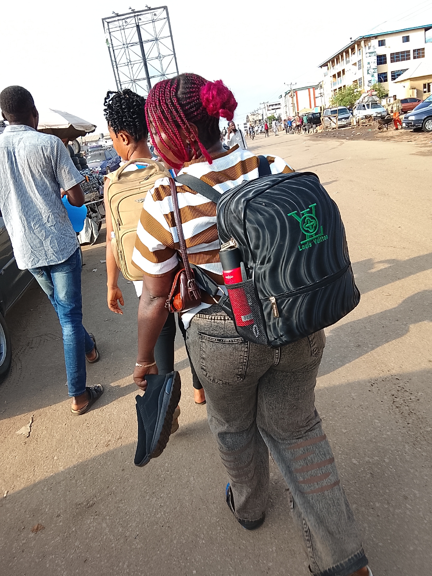 Three people walk along a street, with other individuals and vehicles visible in the background. A man with a light blue shirt and blue jeans is on the left, walking away from the camera. Next to him is a woman with curly black hair, wearing a light brown backpack. To the right, a woman with bright pink braided hair is wearing a brown and white striped shirt, grey jeans with distressed designs, and a black backpack with a "Louis Vuitton" logo. She is holding a black shoe in her hand. The street is paved and appears to be in a somewhat busy urban area. Buildings are visible in the distance. The location is the city of Abuja, Nigeria.