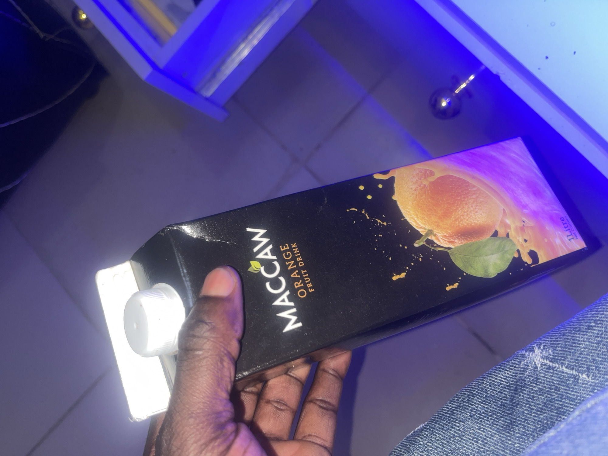 A person's dark-skinned hand is holding a black juice carton, brand name "MACCAW," with an orange graphic. The carton has a white cap and appears to be a 1-liter beverage. The background is dark, with purple and blue lighting. The lighting suggests an indoor setting, possibly a room or a bar, with a blue illuminated structure visible on the right side. The overall lighting conditions are low, with the carton being the primary illuminated subject. There is no discernible information suggesting a specific location within Kabong, Nigeria.