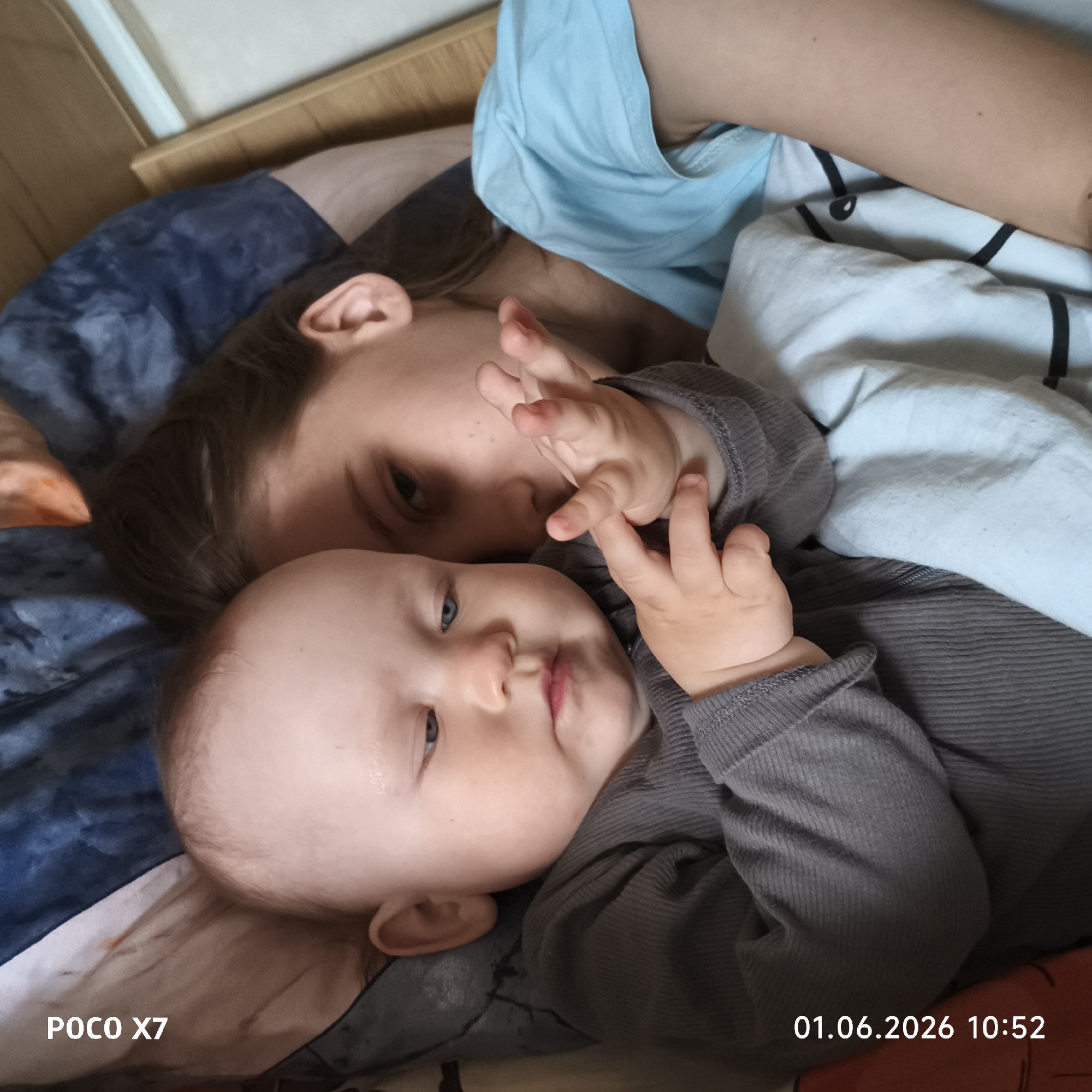 The image depicts a young child and a baby lying on a bed. The child, appearing older, has brown hair and is positioned behind the baby. The baby is in the foreground, wearing a gray long-sleeved shirt and has their hand raised near their face. The bedding is a mix of blue, gray, and white. The environment seems to be an indoor bedroom setting. There is a timestamp of "01.06.2026 10:52" in the bottom right corner and "POCO X7" in the bottom left corner. The location is not discernible from the image.