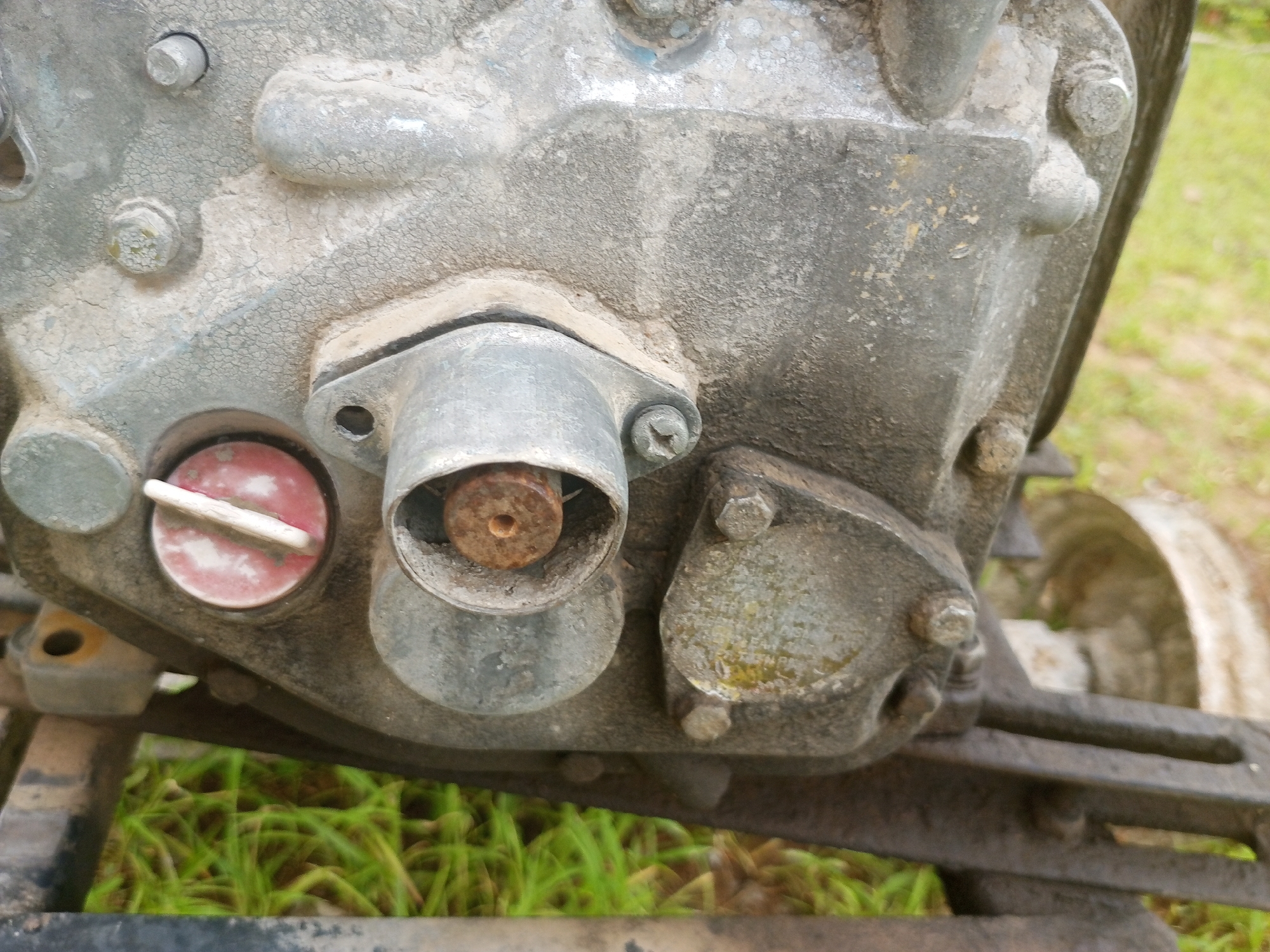 This image shows a close-up view of an old, weathered metal engine, likely from a piece of machinery or a small vehicle. The engine is gray and covered in dirt and grime, with some areas showing cracked paint. It is mounted on a dark metal frame.
Several components are visible on the engine's surface. There are various bolts and screws holding parts together, some of which appear rusted. One prominent feature is a metallic fitting with a round, rusty metal rod protruding from its center, possibly a valve or a part of the engine's internal mechanism. To the left of this fitting, there is a circular cap with a red and white pattern, secured by a metal lever. This cap could be a drain plug or a fill cap. Another smaller, round metallic cover is located to the right of the central fitting, appearing slightly greasy. In the background, a portion of a wheel, also appearing weathered and metallic, is visible, along with a blurred backdrop of green grass.
The scene appears to be outdoors, suggested by the natural lighting and the presence of grass. The time of day is difficult to determine precisely, but the lighting suggests it could be daytime. There are no people visible in the image. The overall impression is one of an aged piece of equipment that has seen significant use and is perhaps no longer in active service, given its worn appearance and the dirt accumulation. There is no discernible text visible in the image. The setting is likely a rural or outdoor industrial area, with the location context suggesting Jalingo, Nigeria.