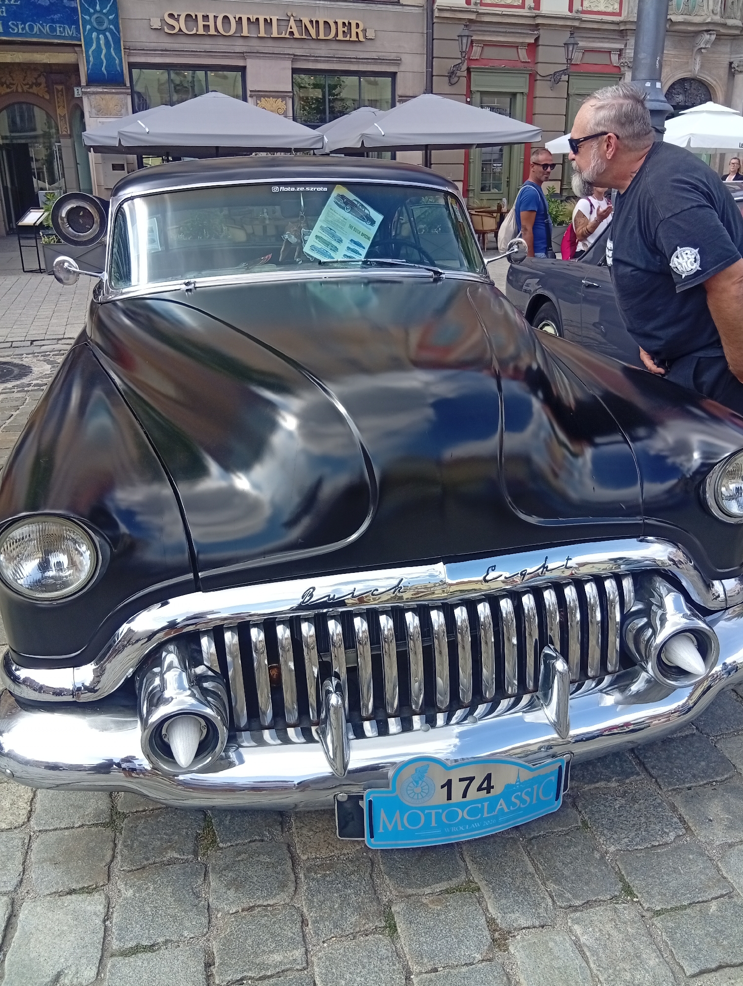 A mid-shot photograph captures a black vintage Buick Eight parked on a cobblestone street in Wrocław, Poland. The car's shiny chrome grille and headlights dominate the foreground. A license plate with the text "174 MOTOCLASSIC WROCŁAW 2026" is affixed to the front bumper. In the background, a building with the name "SCHOTTLÄNDER" is visible, suggesting a commercial establishment. Several people are present in the scene; a man with sunglasses and a beard is leaning over the car, looking at it, while other individuals are seen in the background, some standing near umbrellas and other parked vehicles. The overall ambiance suggests a classic car event or exhibition.