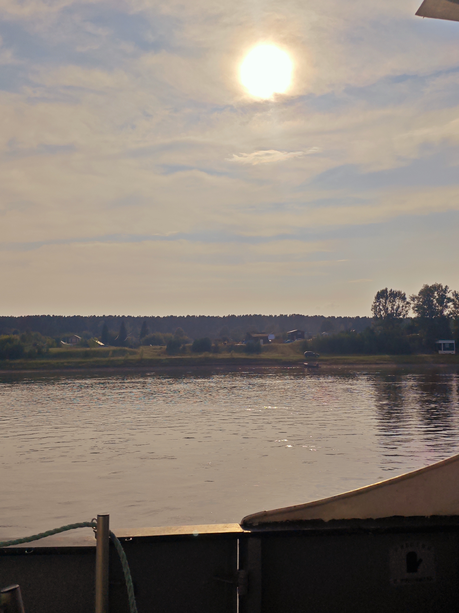 The image depicts a wide body of water reflecting the sky and the sun. The sun, a bright orb, is visible in the upper portion of the sky, partially obscured by soft clouds. On the far bank of the water, a line of trees forms a dense, dark border. In front of the trees, there are scattered small buildings and structures, suggesting a rural settlement. A few taller trees stand out amongst the foliage. In the foreground, the edge of a vessel is visible, with a metal railing and a green rope. The water's surface is rippled, with highlights from the sunlight. The scene appears to be set in the village of Yukseevo, Russia, given the presence of a natural landscape and likely a river or lake. There are no people or overt actions depicted. The environment is tranquil and natural, with a hint of human habitation on the horizon.