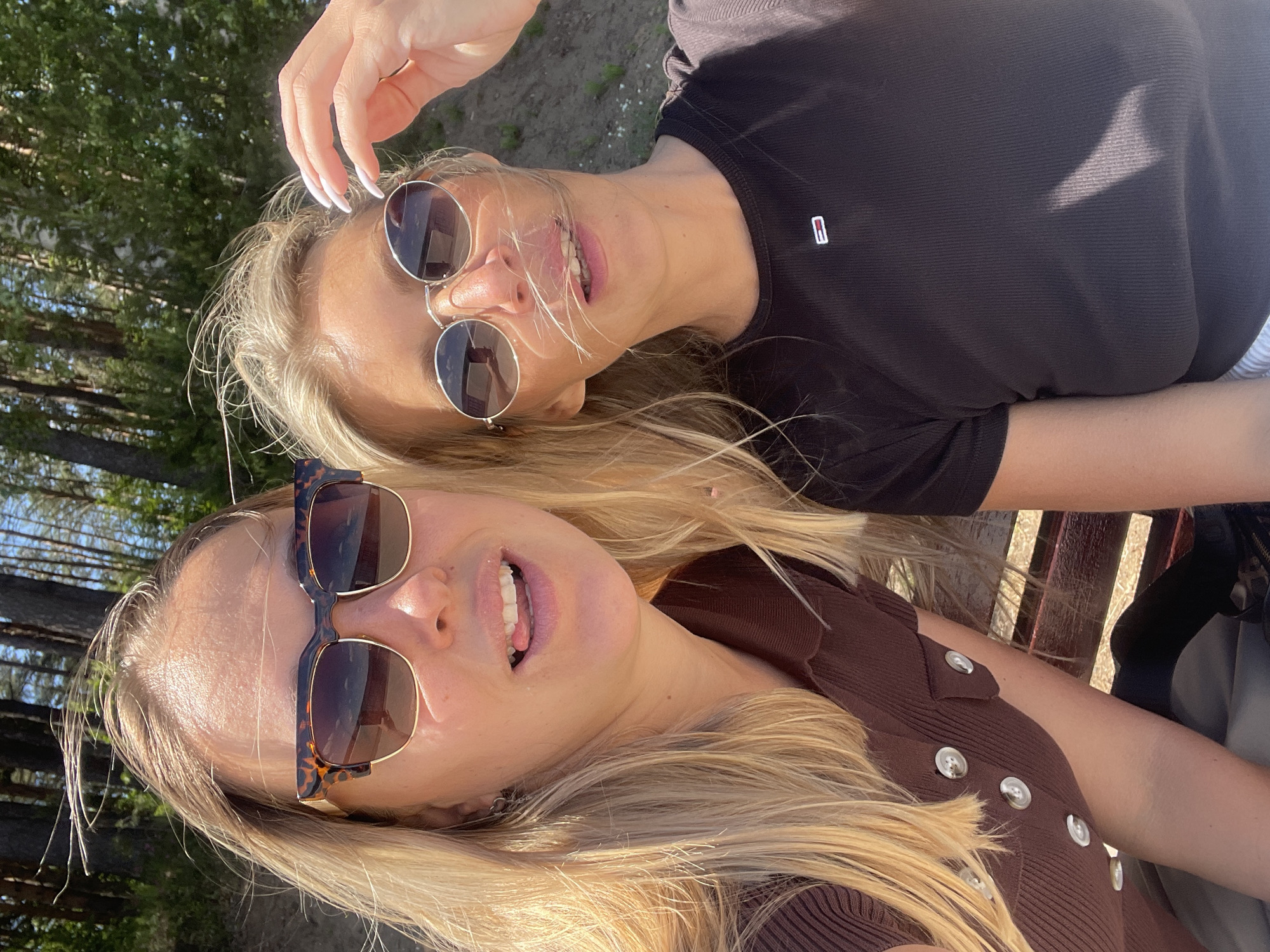 Two women are taking a selfie outdoors in a wooded, sandy area with a slight incline in the background. The woman on the left, with blonde hair, wears a brown button-up top and tortoise-shell sunglasses. She is sticking her tongue out slightly. The woman on the right, also with blonde hair, wears a black t-shirt and round sunglasses. Her hand is raised to touch her hair. The environment suggests a park or beach setting. There is no visual information indicating the location is in Chaikovsky, Russia.