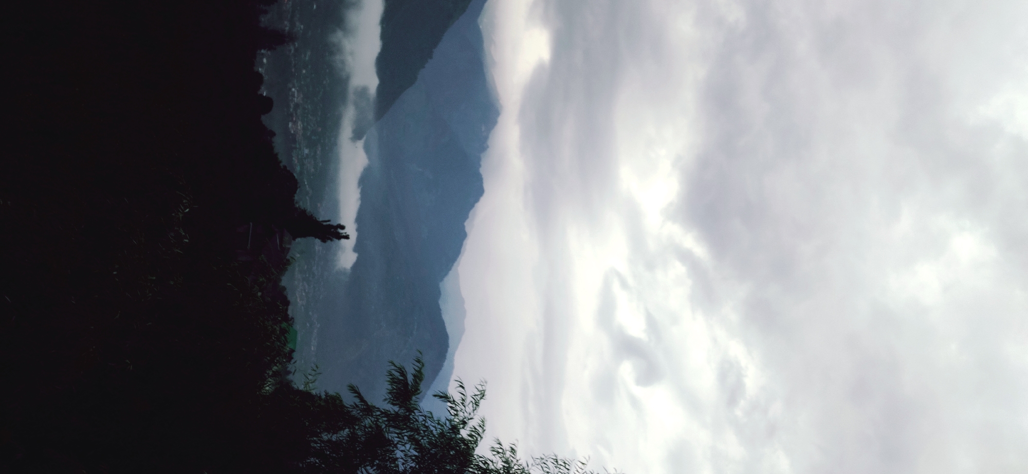 The image depicts a panoramic view of a mountainous landscape with a town nestled in the valley, likely Patar, India. The sky is overcast with dramatic, swirling clouds, suggesting an impending storm or recent rainfall. Layers of mist or low-lying clouds partially obscure the lower slopes of the mountains, creating a sense of depth and mystery. In the foreground, silhouetted trees and foliage frame the scene, adding a dark, almost ominous contrast to the lighter hues of the sky and mountains. The town below is barely visible, its structures appearing as a scattering of small lights and shapes against the darkened terrain. A lone, slender tree with a distinctive silhouette stands prominently in the mid-ground, adding a touch of verticality to the horizontal expanse of the mountains and valley. The overall mood is one of serene, yet powerful, natural beauty, with a hint of the dramatic weather that may be present in the region.
